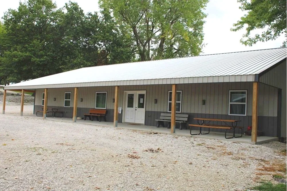 Single-story building with a metal roof, beige siding, several windows, and a white door, surrounded by trees and gravel ground. Big porch with picnic tables