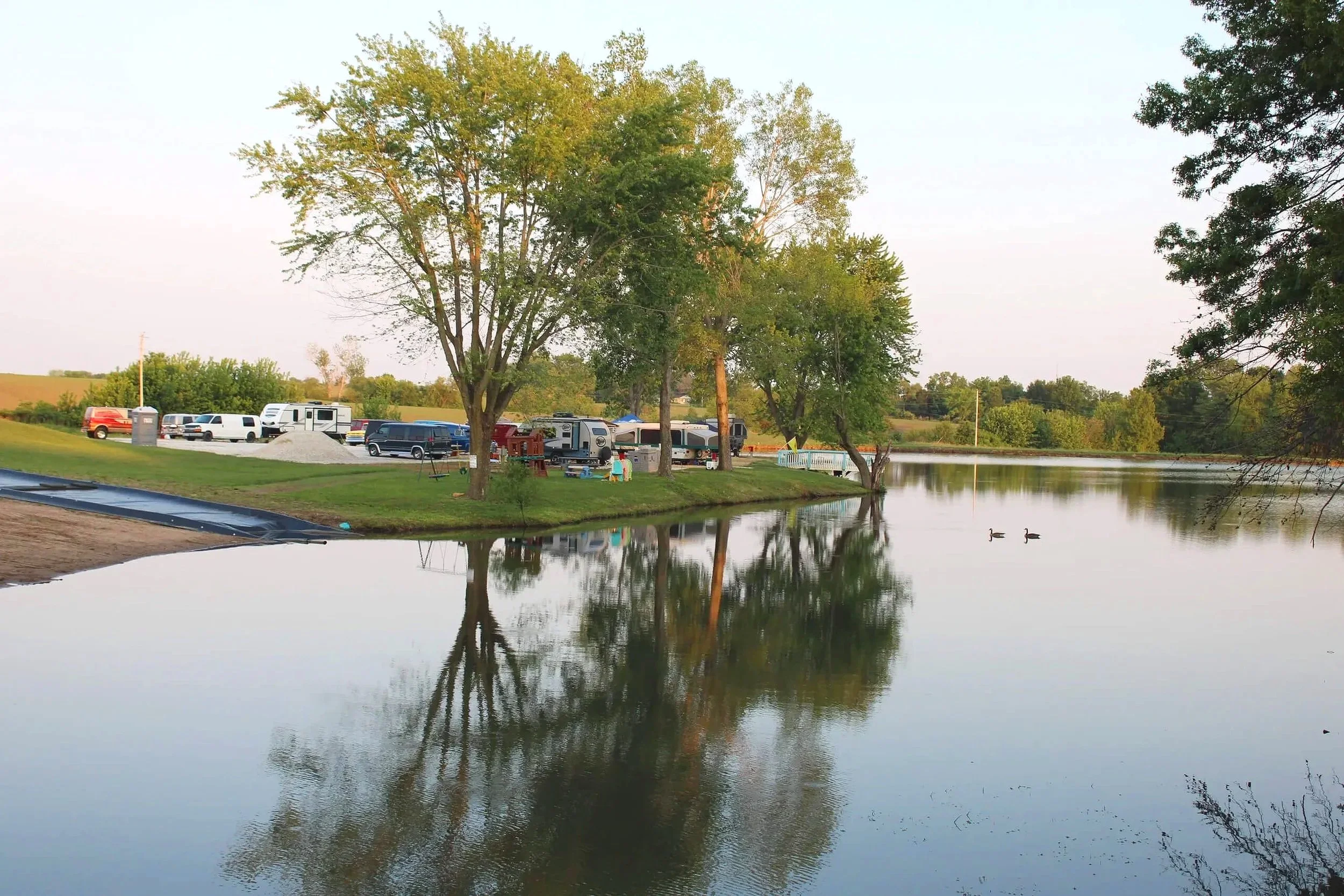 A peaceful lakeside scene with trees, parked recreational vehicles, and ducks swimming in the water, during sunset.
