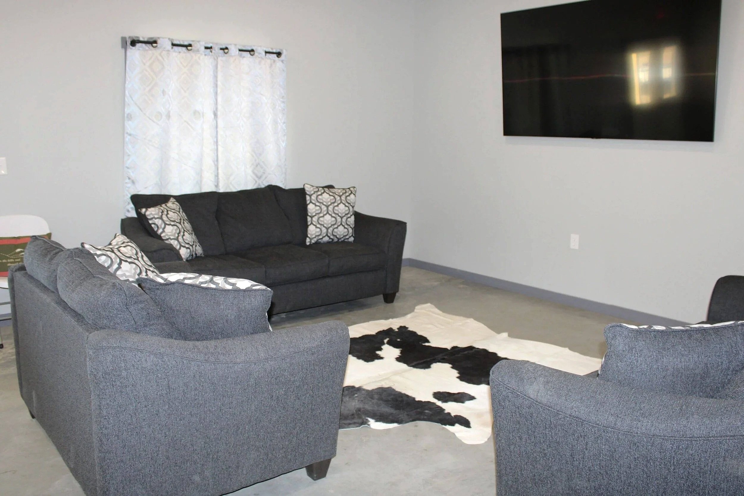 Living room with a gray couch and armchair, black and white patterned pillows, a black and white cowhide rug, light-colored walls, a window with white patterned curtains, and a large flat-screen TV mounted on the wall.