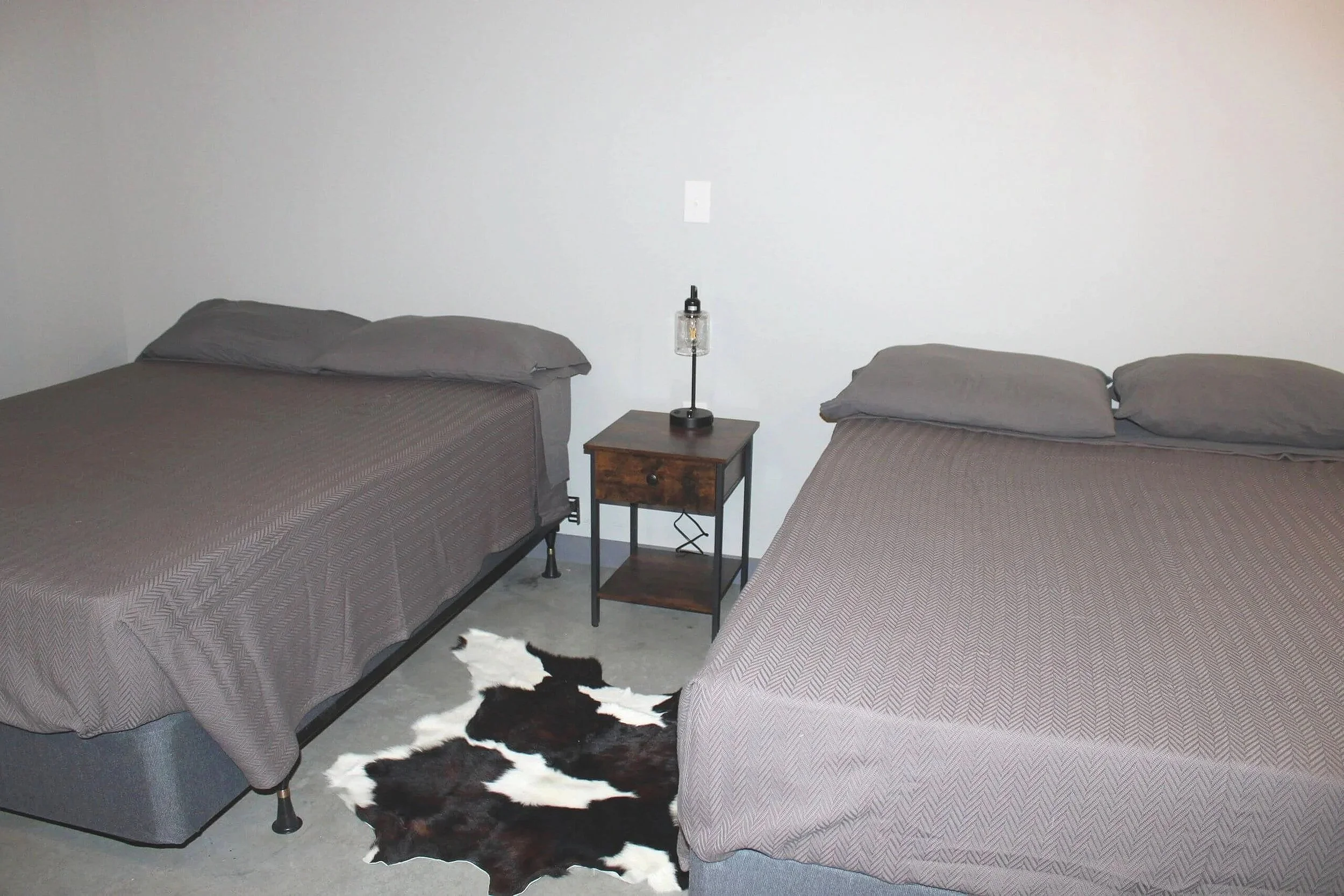 A bedroom with two beds with gray bedspreads and matching pillows. A nightstand with a lamp between the beds. A cowhide rug on the floor near the foot of the beds. A plain white wall in the background with an electrical outlet.