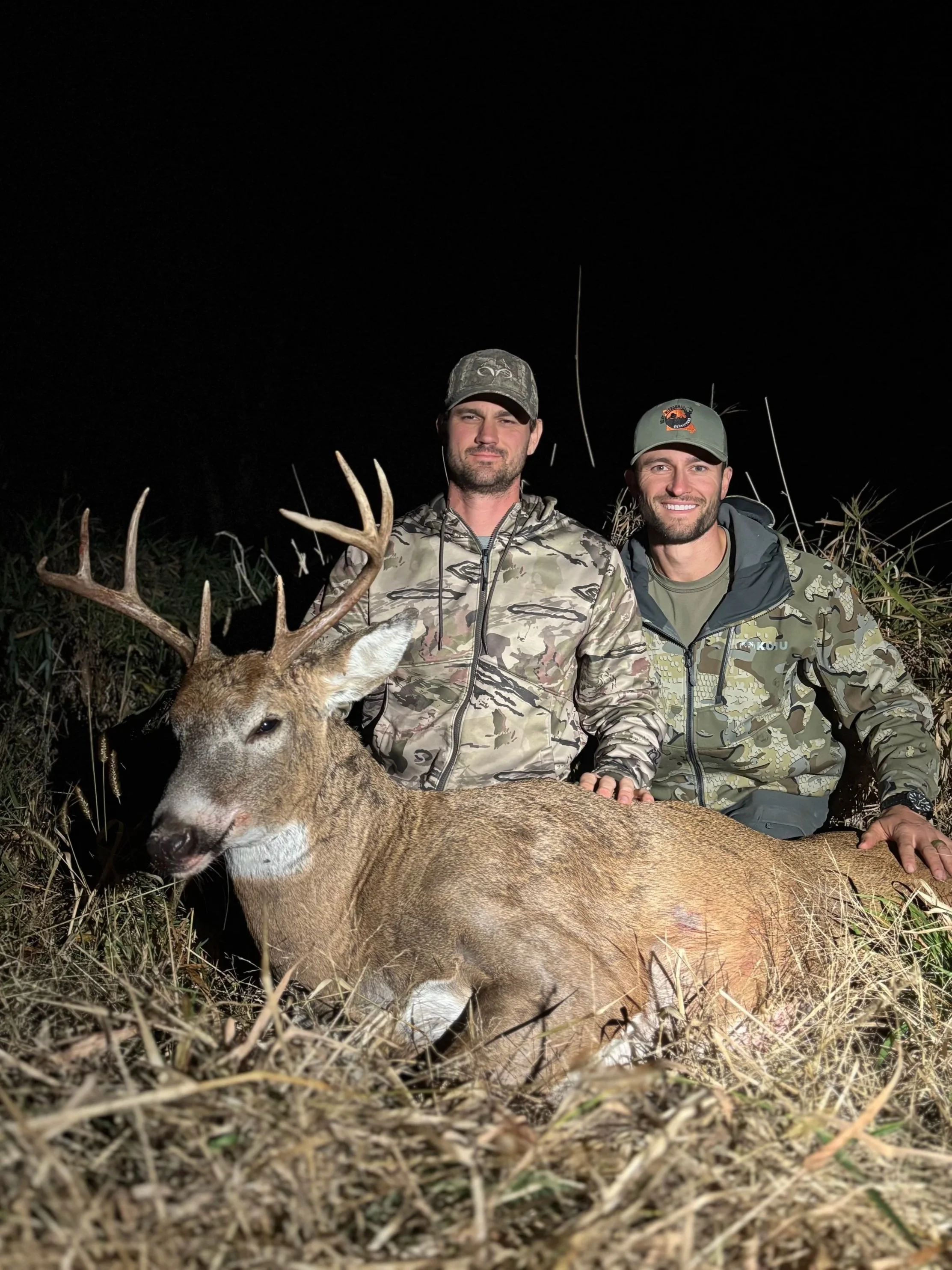 Two men in camouflage jackets and hats sitting next to a large deer they have hunted, with the deer's head and antlers in the foreground, against a dark night background.