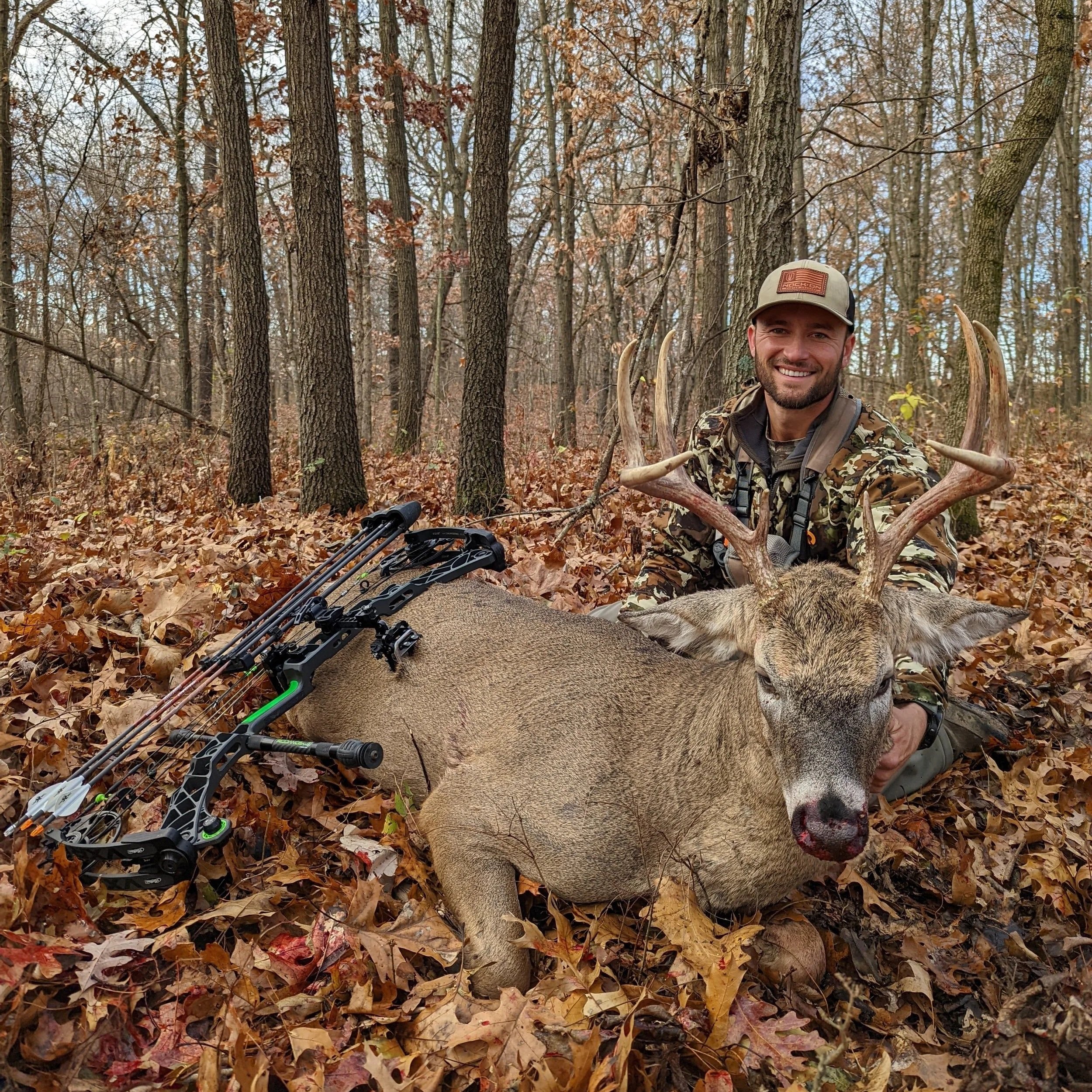 A man in camouflage clothing and a beige cap smiling in a forest during autumn, with a fallen deer lying on the ground in front of him, and a compound bow placed on the deer's side.