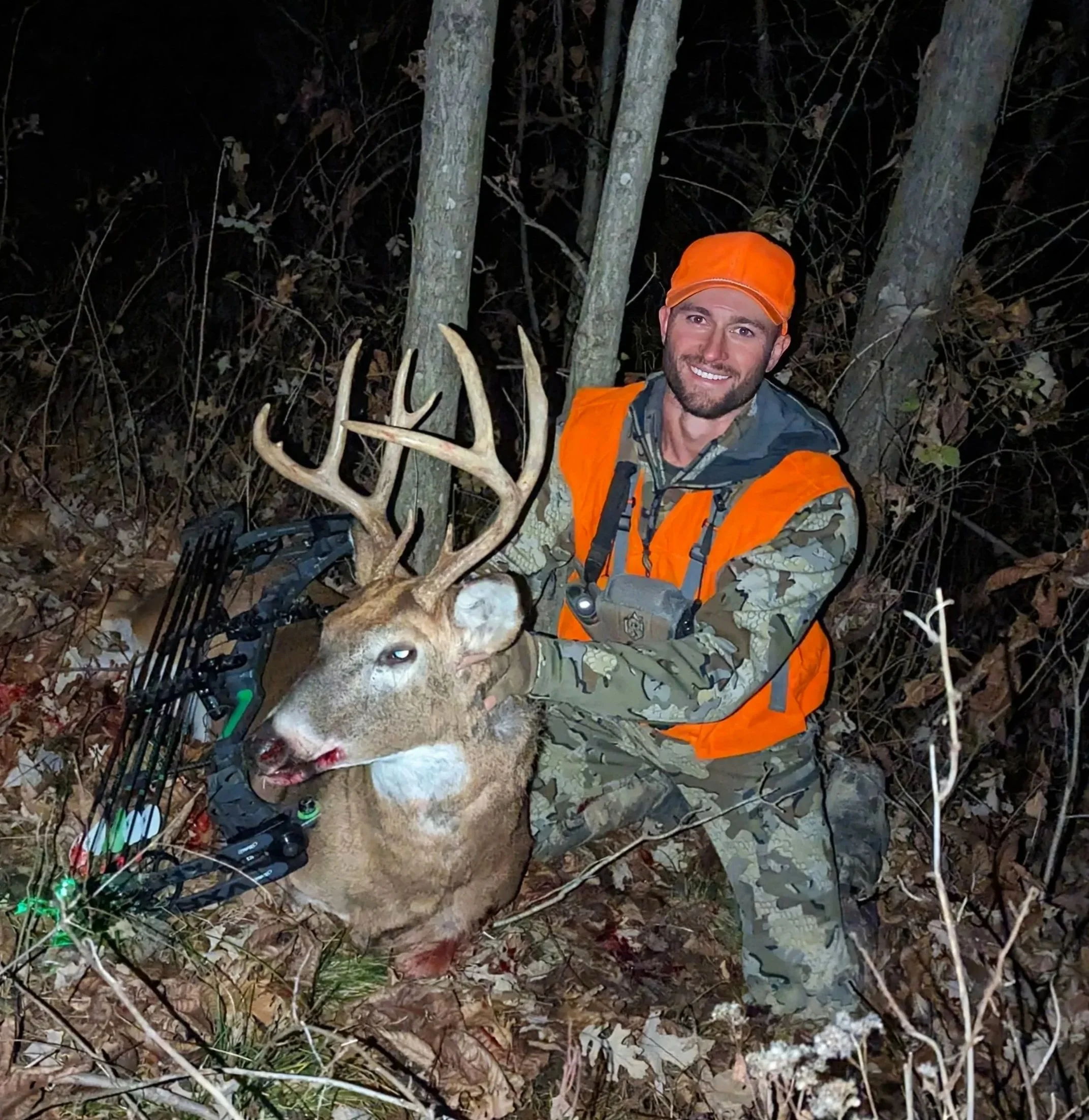 A man wearing an orange hunting vest and hat kneels in a wooded area at night, posing with a large deer he has hunted, which has prominent antlers. There is a compound bow leaning against its side