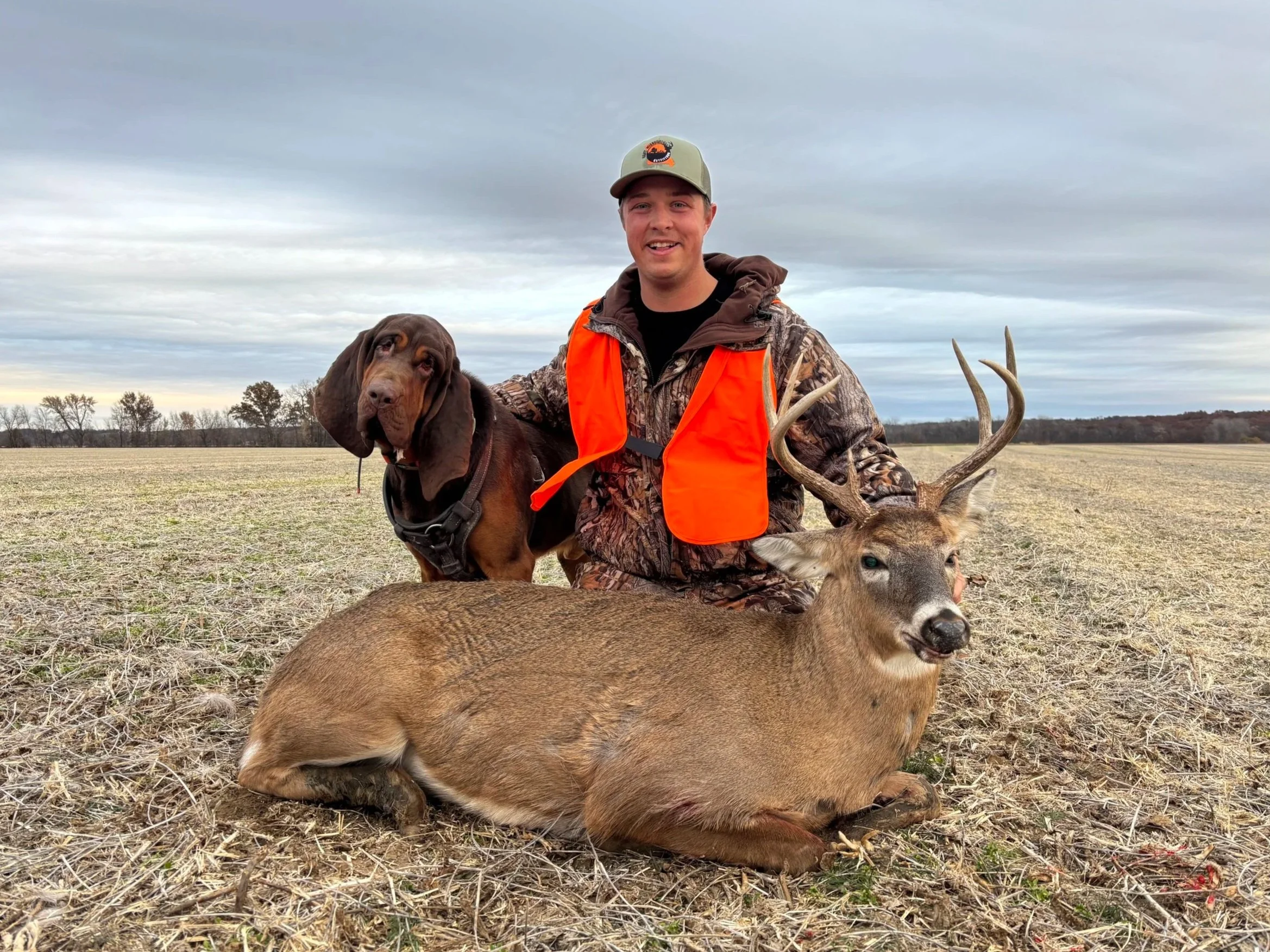 A man in camouflage clothing with an orange vest and cap, holding a deer with antlers and a bloodhound dog, in an open field with cloudy sky.