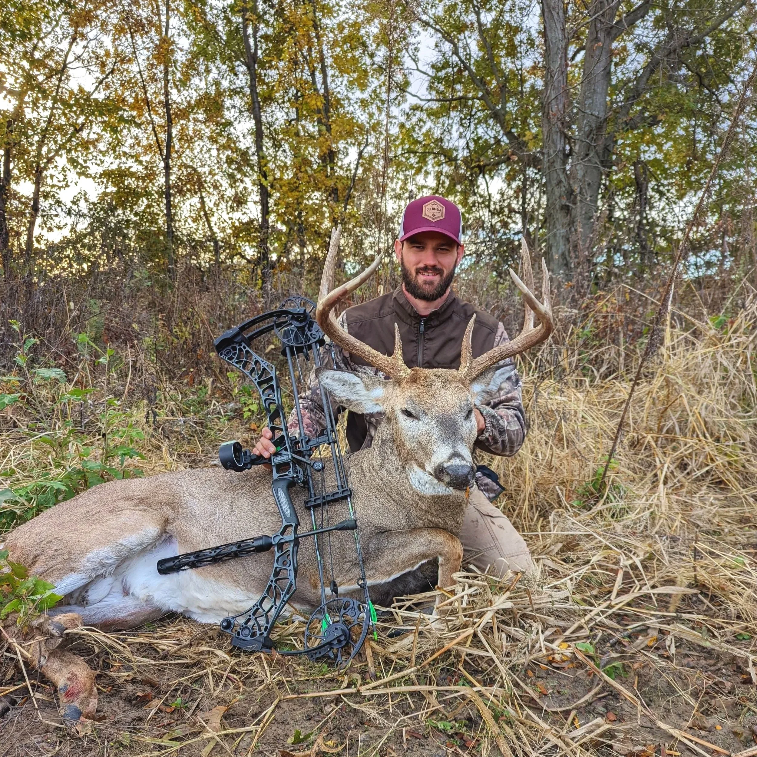 A man in outdoor camouflage clothing and a maroon hat holding a deer with a bow and arrow in a wooded area with trees and foliage.