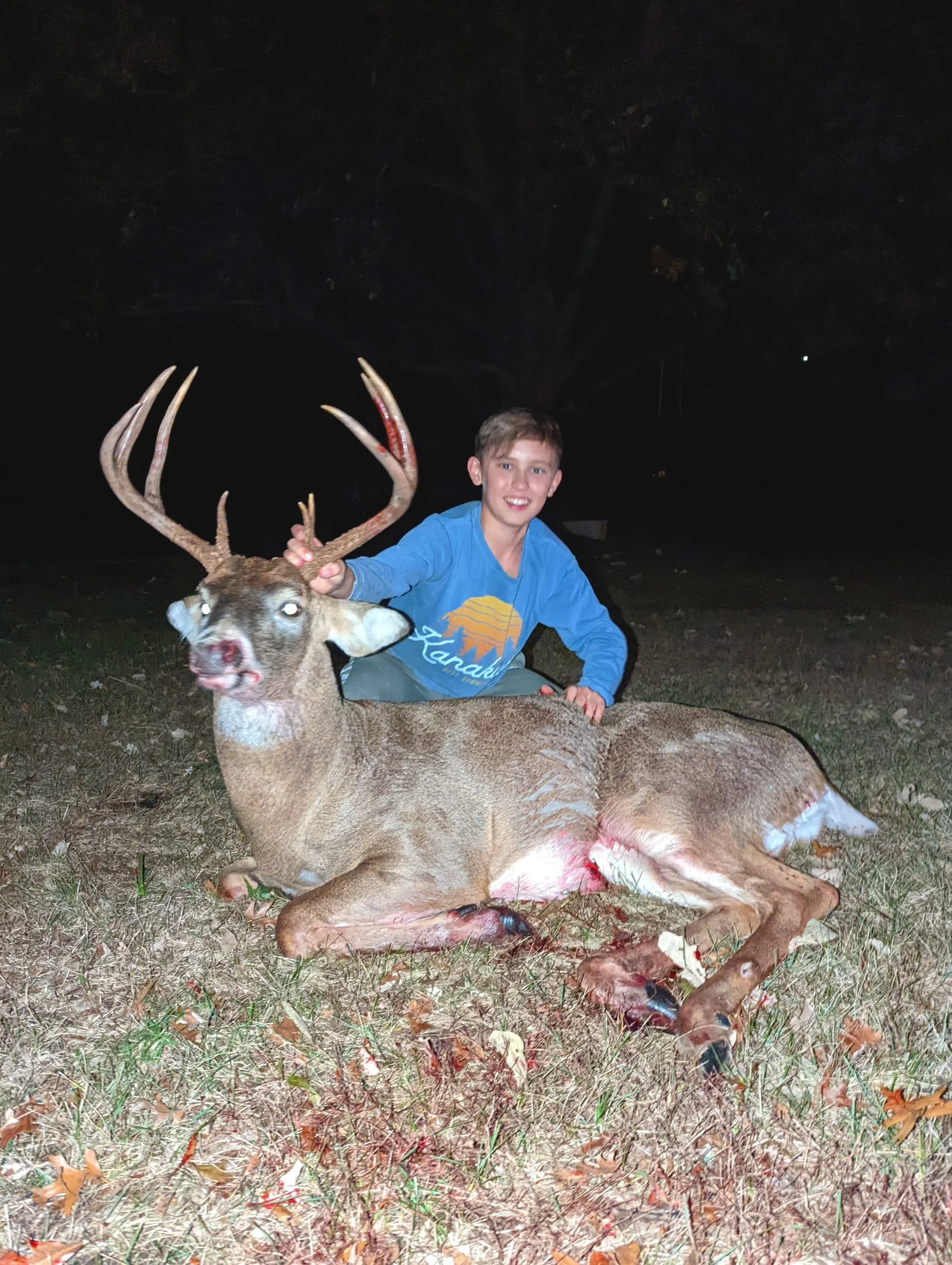 A young boy in a blue shirt kneels next to a dead deer with large antlers, marked with blood, lying on the grass at night.