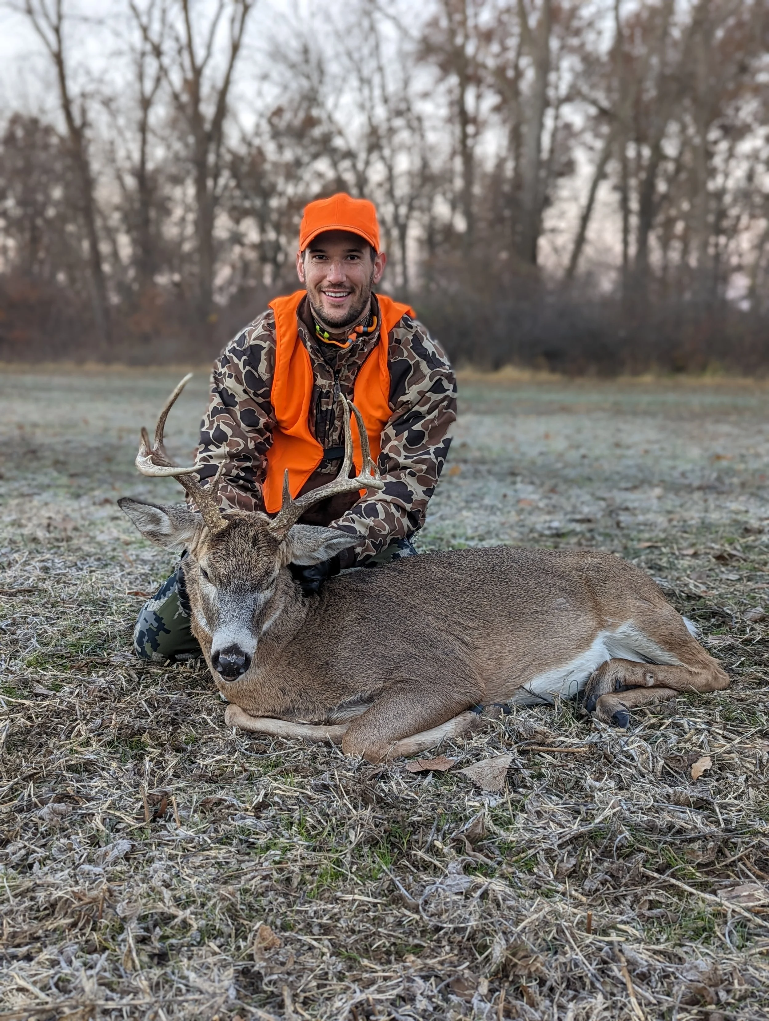 A man in outdoor hunting clothes kneels next to a large deer with antlers, lying on the ground in a wooded area during late fall or winter.