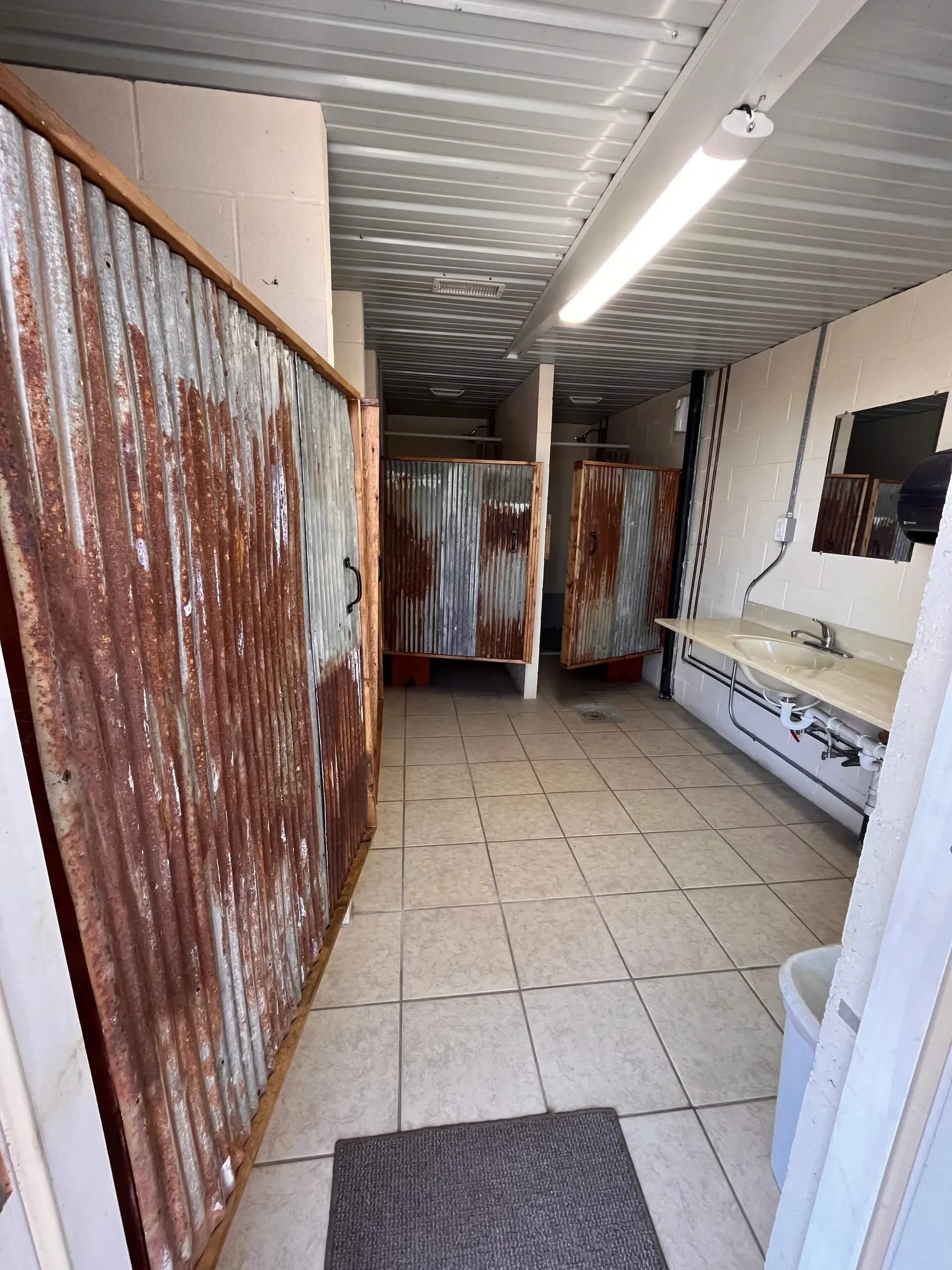 Public restroom with wooden and metal stalls, tiled floor, sink with faucet, and fluorescent lighting.