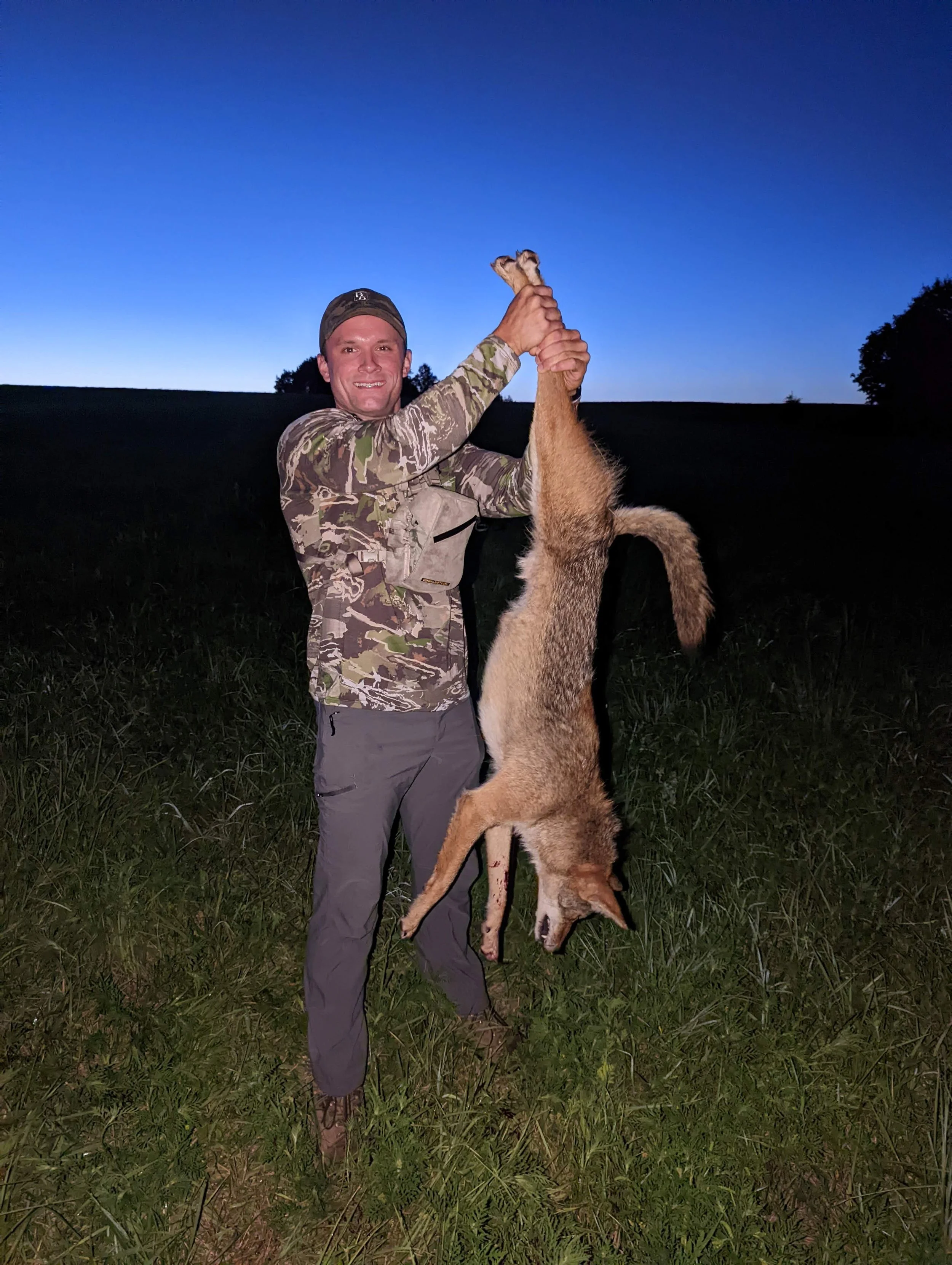 A smiling man in camouflage clothing holding a large coyote by its front paws, with the coyote upside down in a grassy field during dusk, with a darkening sky and some trees in the background.