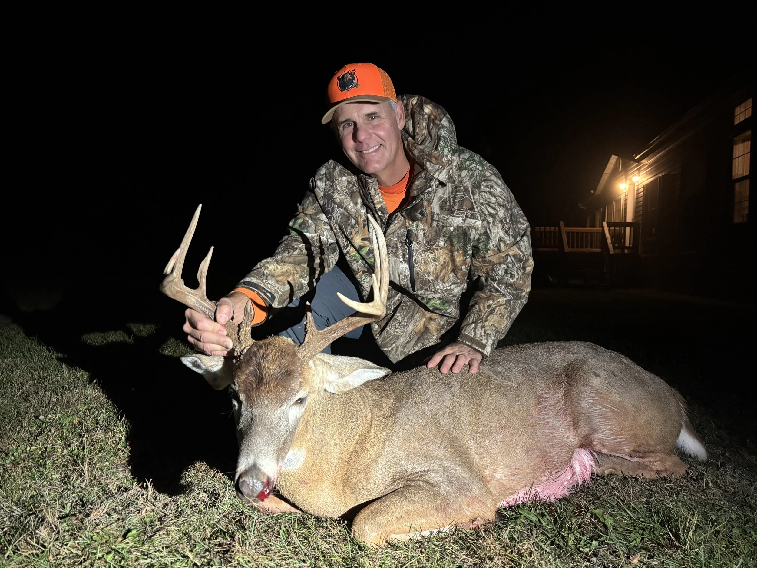 A man in camouflage clothing and an orange cap is smiling while holding an antlered deer carcass at night. There is a house with lights on in the background.