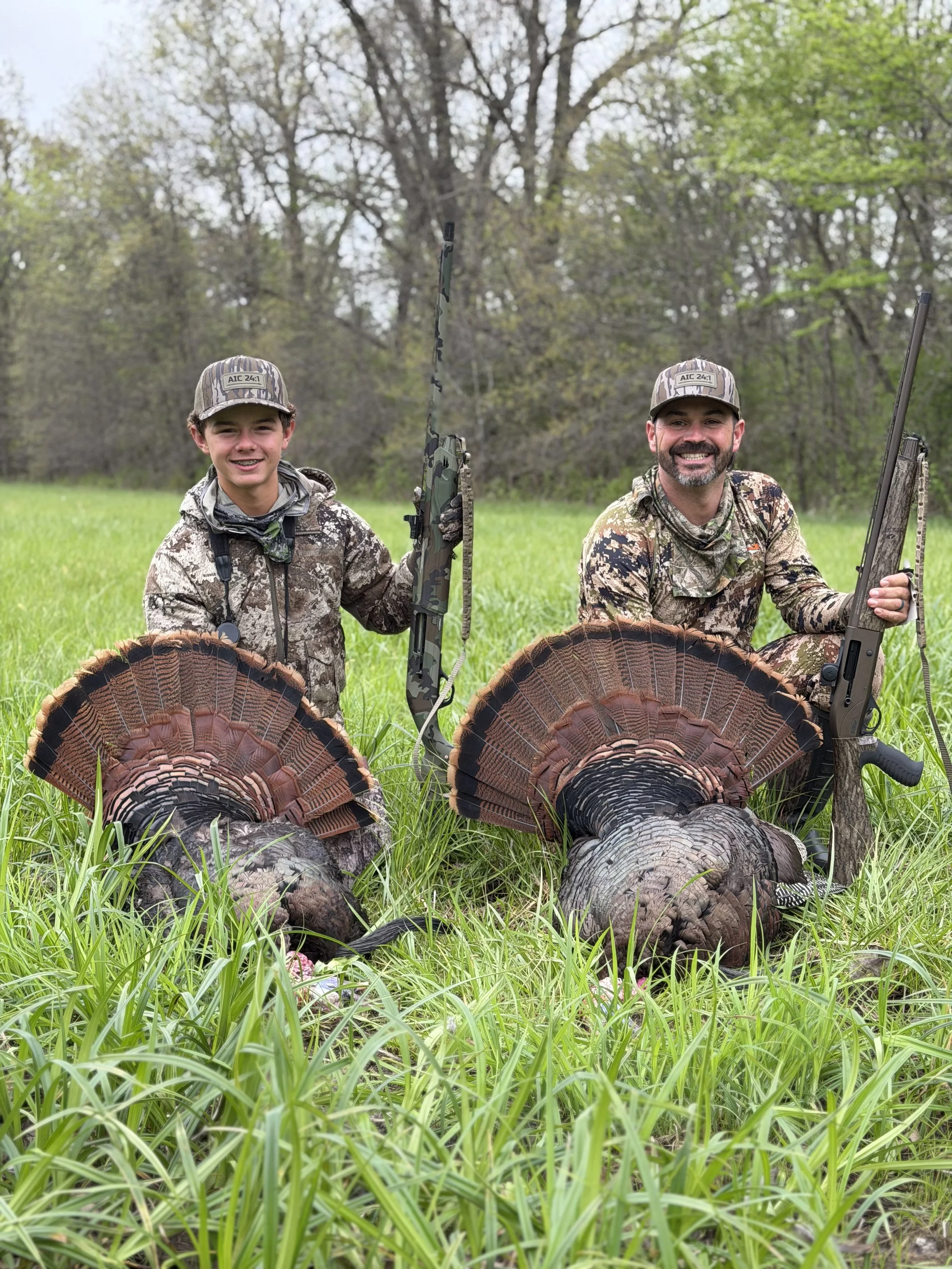 Two men dressed in camouflage hunting attire kneel beside two large wild turkeys with fanned tail feathers on a grassy field in front of a background of trees holding shotguns.