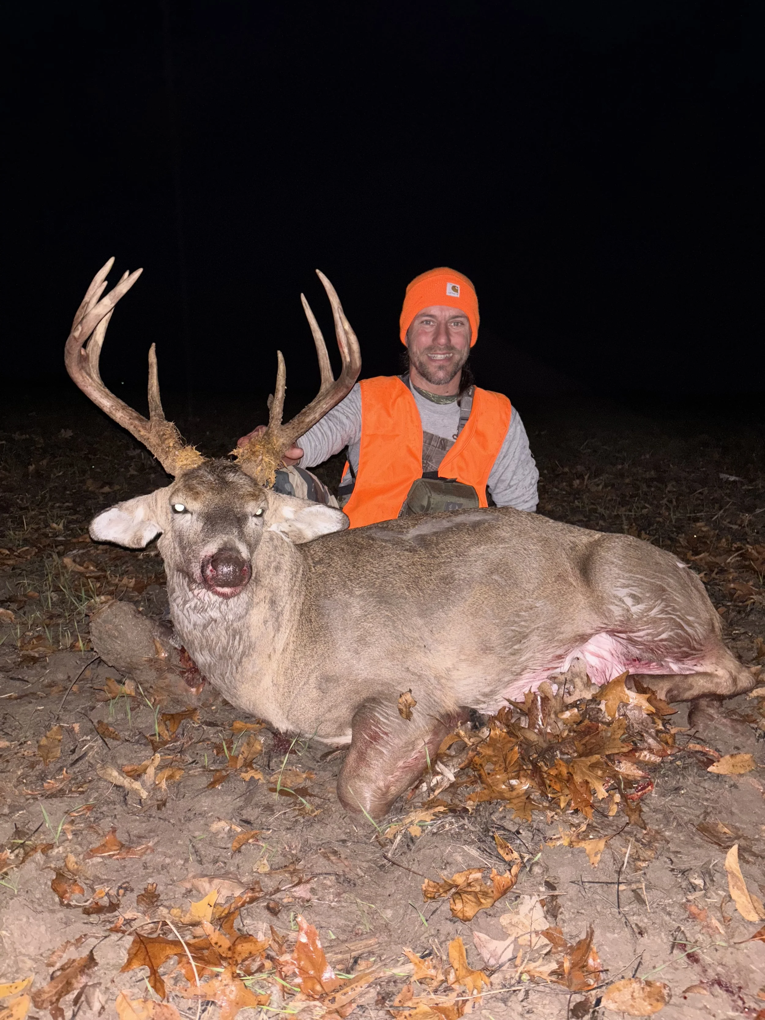 A man dressed in hunting gear, wearing an orange hat and vest, kneeling next to a large, recently hunted deer with antlers, lying on the ground among fallen leaves at night.