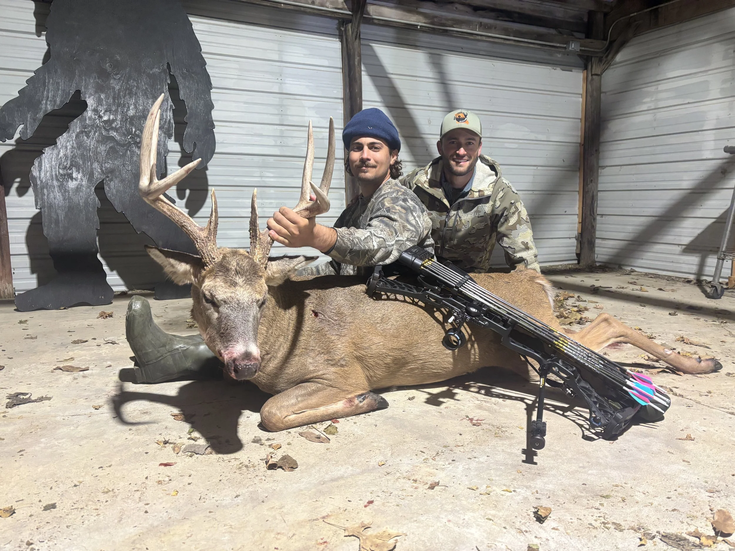 Two men in camouflage clothing pose with a deer and a bow, inside a shed with metal walls, and a silhouette of a big foot decoration on the wall. One man holds the deer's antlers, and the other sits behind.