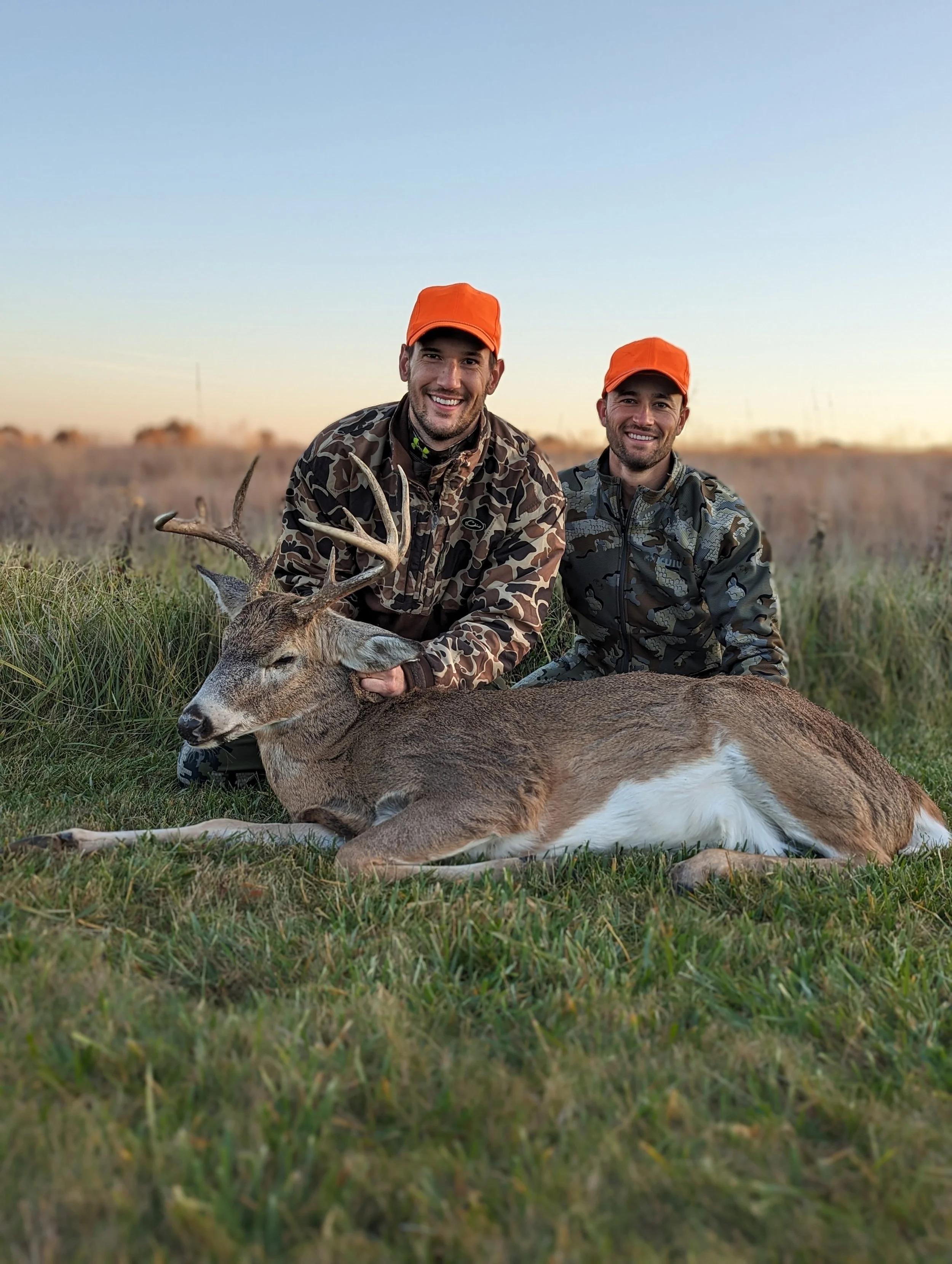 Two smiling men in camouflage jackets and orange caps sitting on grass with a dead deer in front of them during sunset in a field.