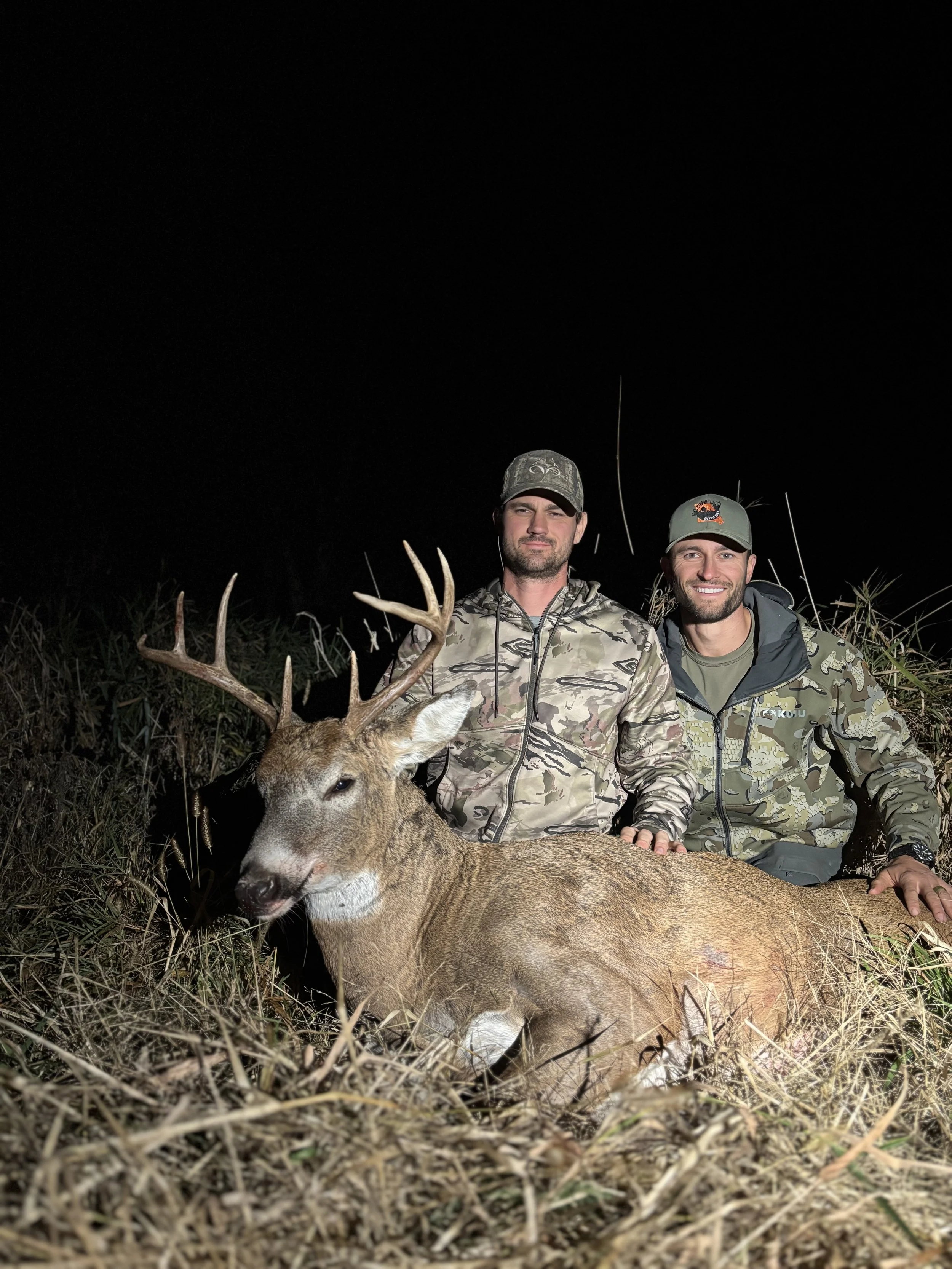 Two men wearing camouflage clothing and caps kneel next to a dead deer with large antlers, outdoors at night.
