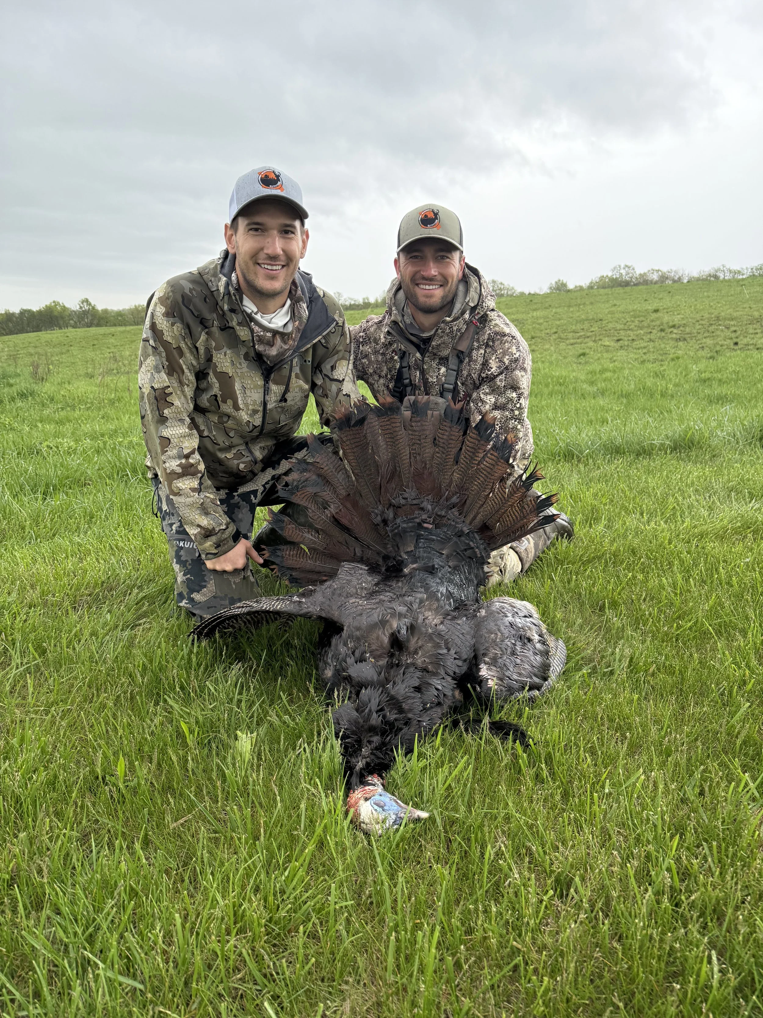 Two men in camouflage clothing and hats smiling and kneeling on green grass in an open field with cloudy sky. They are posing with a dead turkey laid out on the grass.