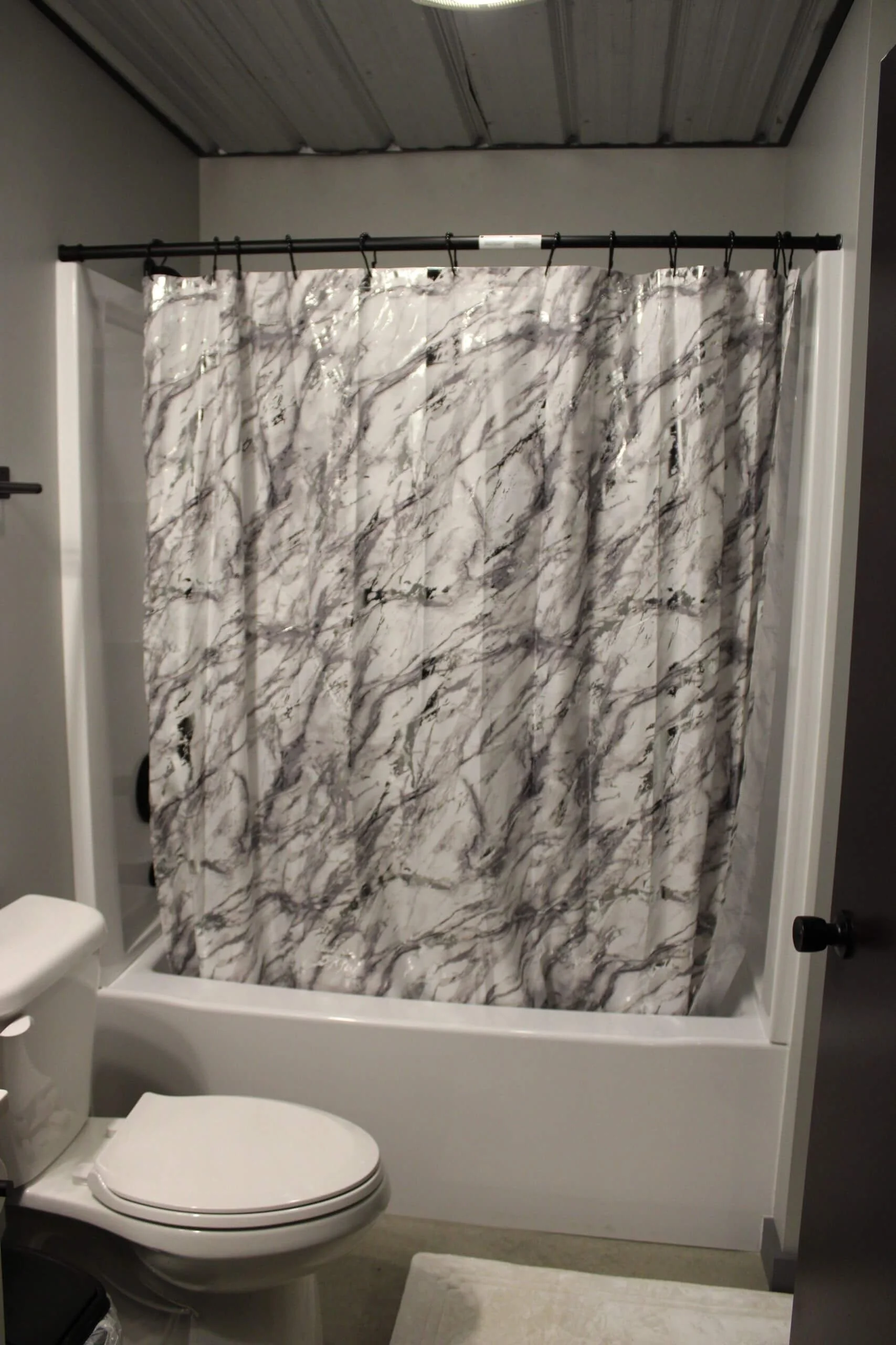 Bathroom with a toilet, a shower with a marble-patterned shower curtain, a partial view of a white toilet, a light-colored bathmat, and a gray wall.