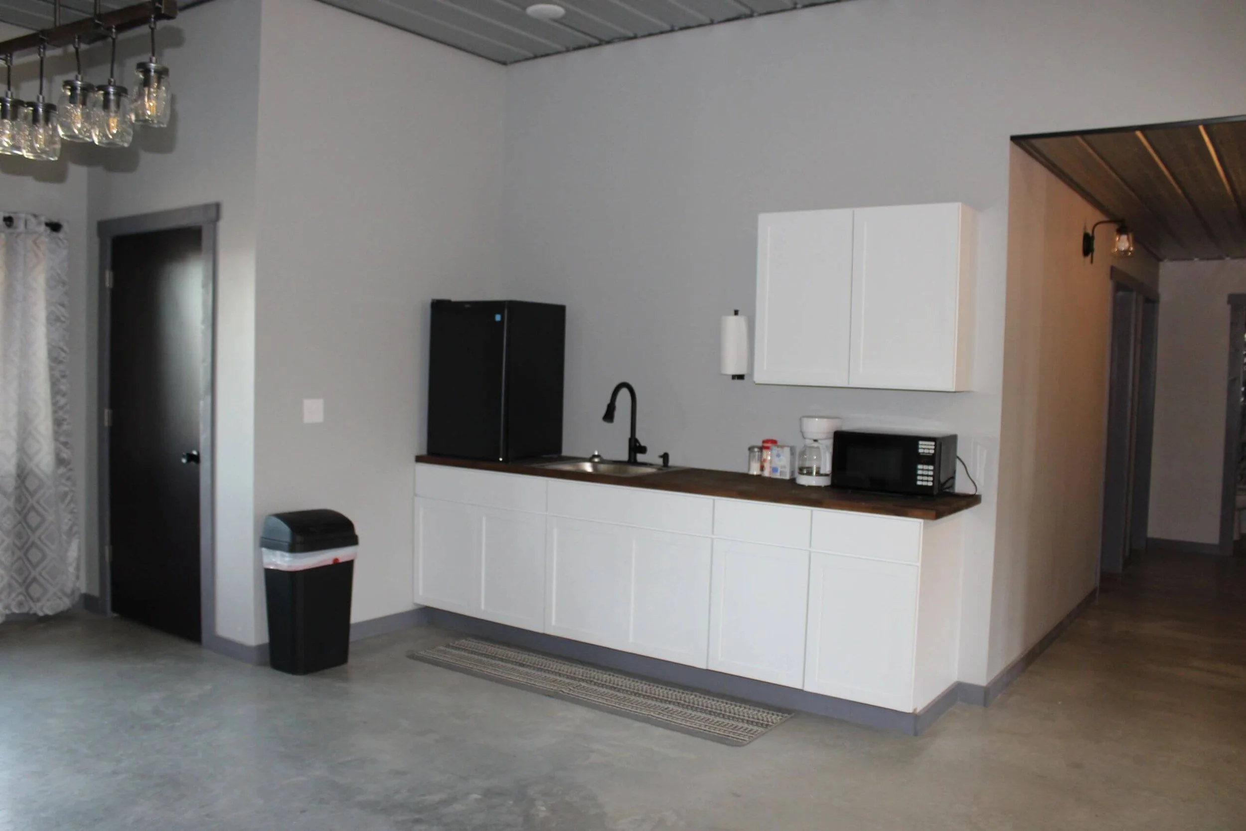 Contemporary kitchen with white cabinets, black fridge, microwave, and black faucet. There are clear pendant lights hanging from the ceiling, and a window with patterned curtains. The room has hardwood flooring and a gray wall.