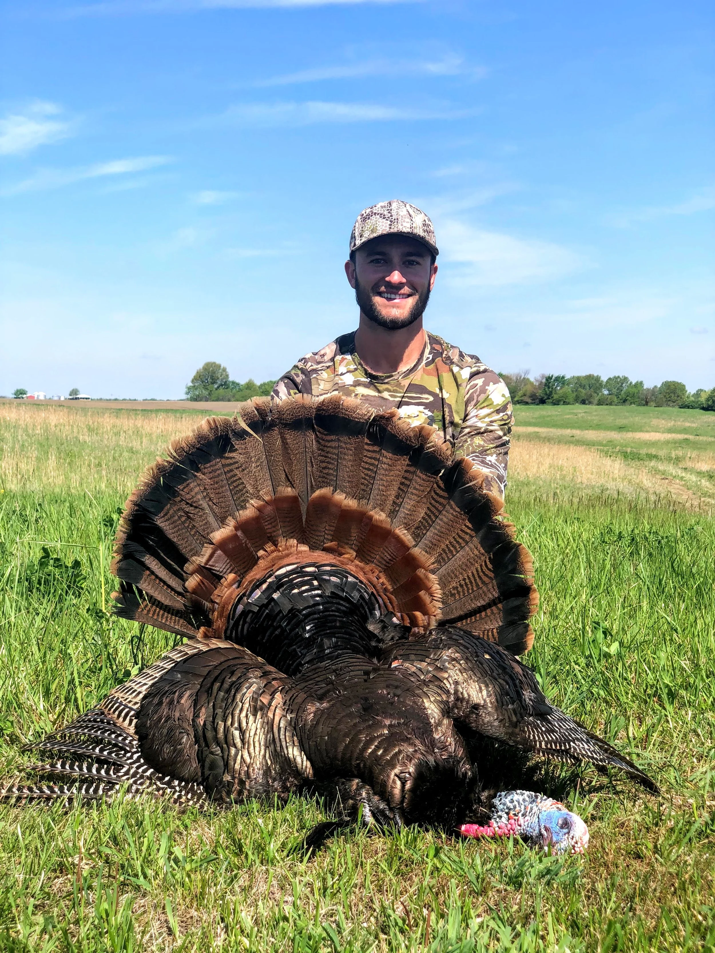 A man in camouflage clothing and a camouflage hat holding a large turkey in a grassy field under a blue sky with some clouds.