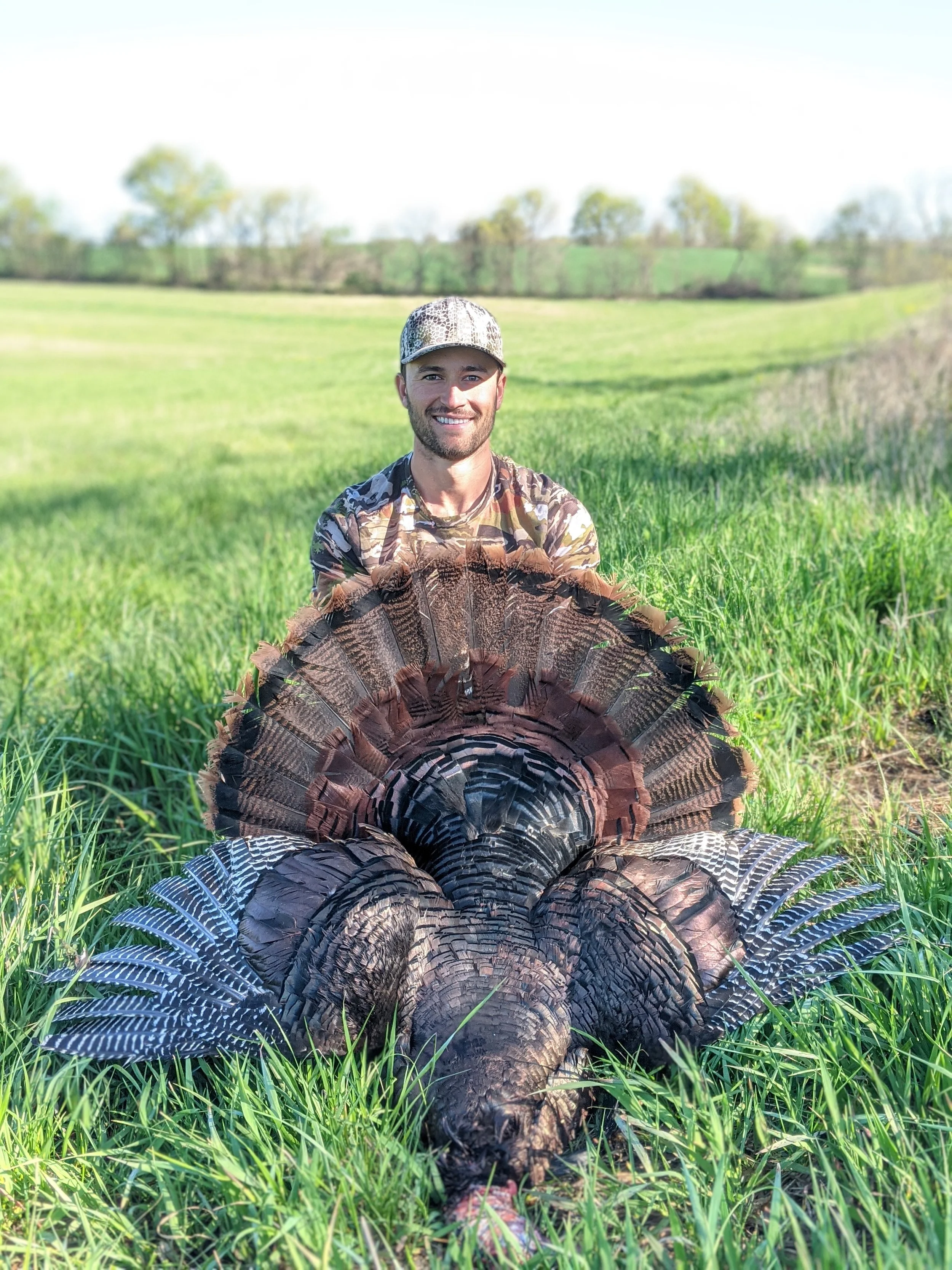A man sitting in a field of green grass holding the fan of a large wild turkey with its tail fanned out behind him, in a rural landscape with trees in the background.