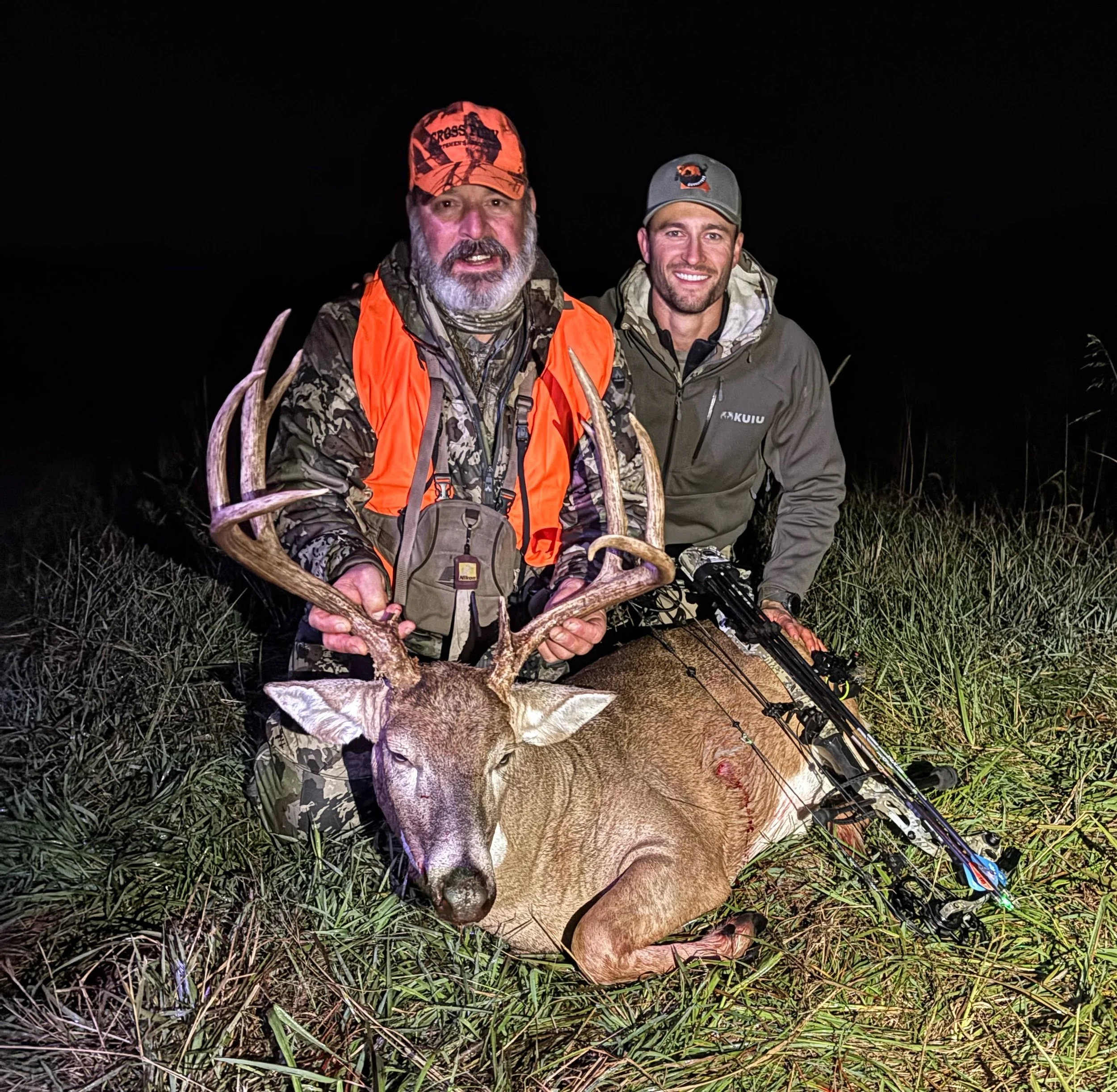 Two men dressed in camouflage and outdoor gear at night, holding a large deer with antlers and a bow and arrow nearby.