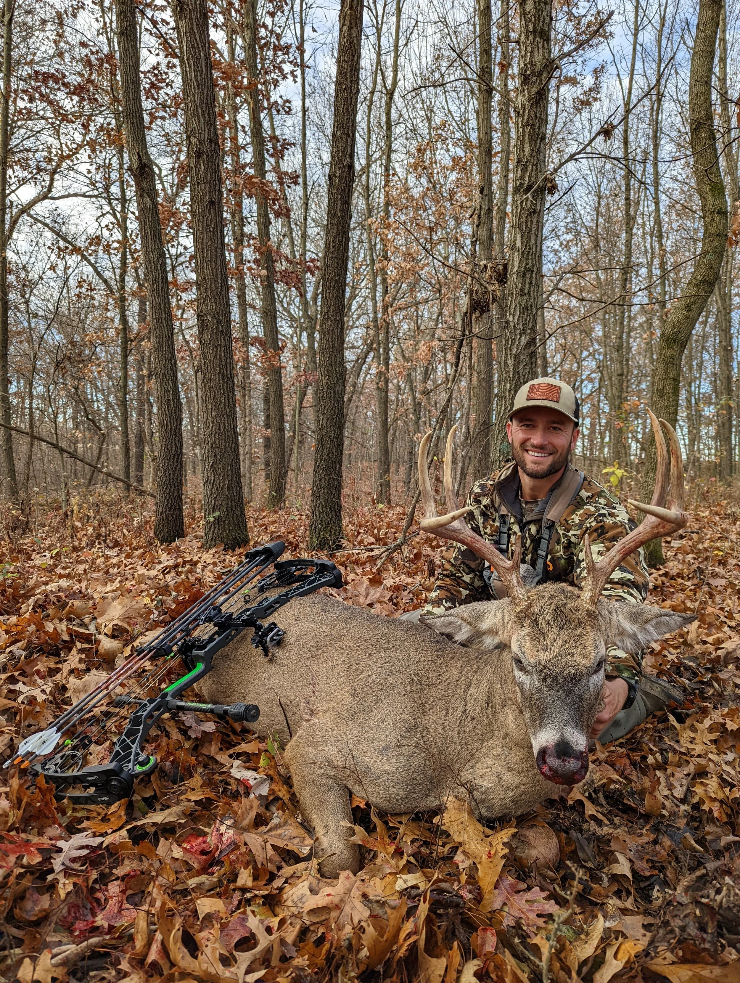 A smiling man in camouflage hunting gear kneels in a wooded forest, holding the corpse of a large deer with antlers. The deer is lying on the ground among fallen leaves, and a bow is resting on the forest floor beside it.