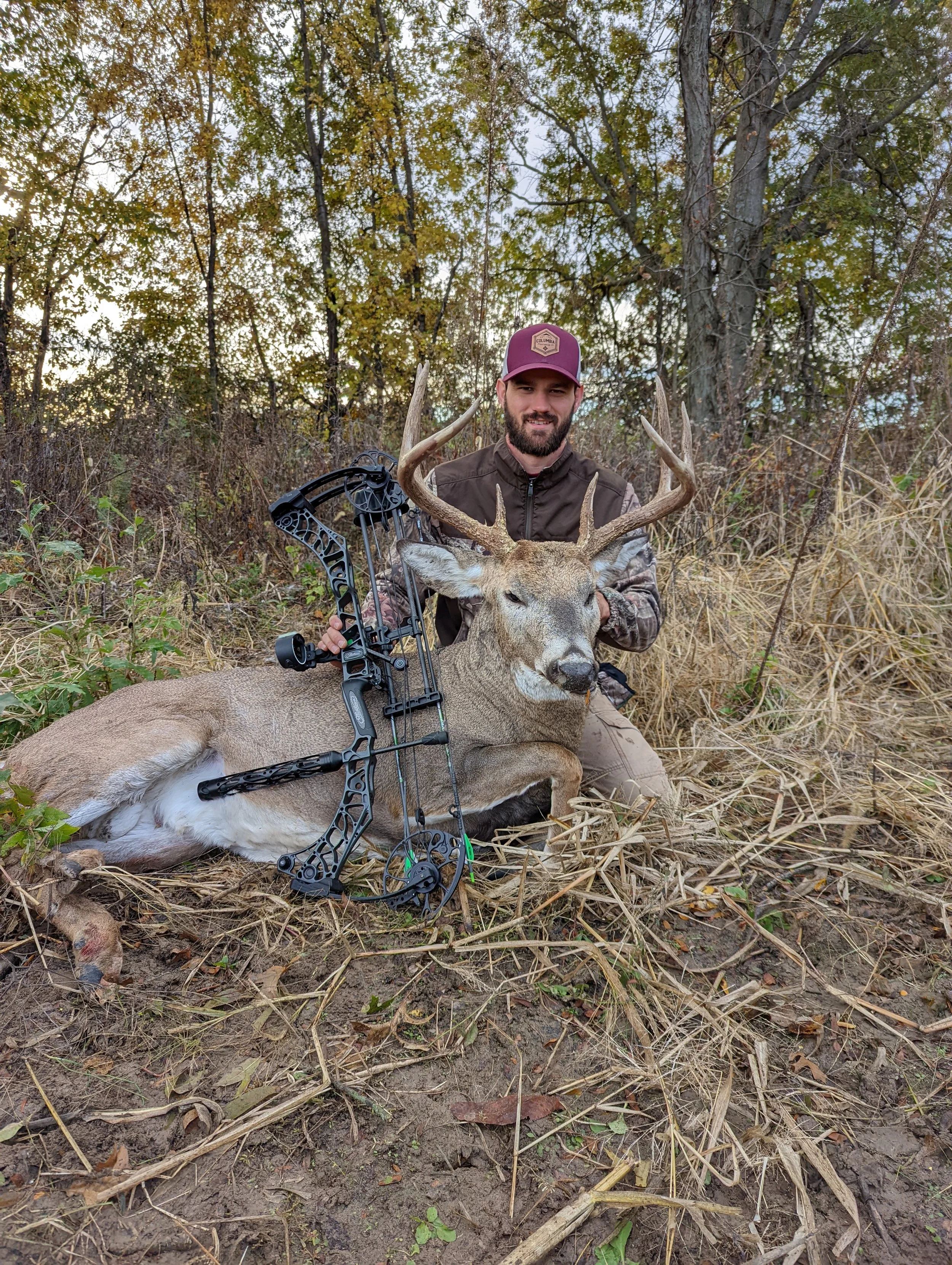 A man sitting on the ground in a forested area, holding a large buck with prominent antlers. A compound bow is resting on the deer, and the man is dressed in camouflage and a maroon cap.