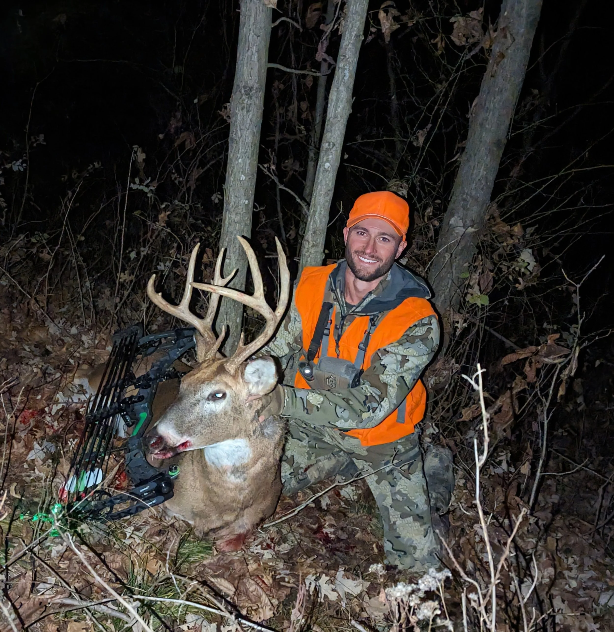 A man wearing an orange hat and jacket in a wooded area at night, holding a large deer with prominent antlers.