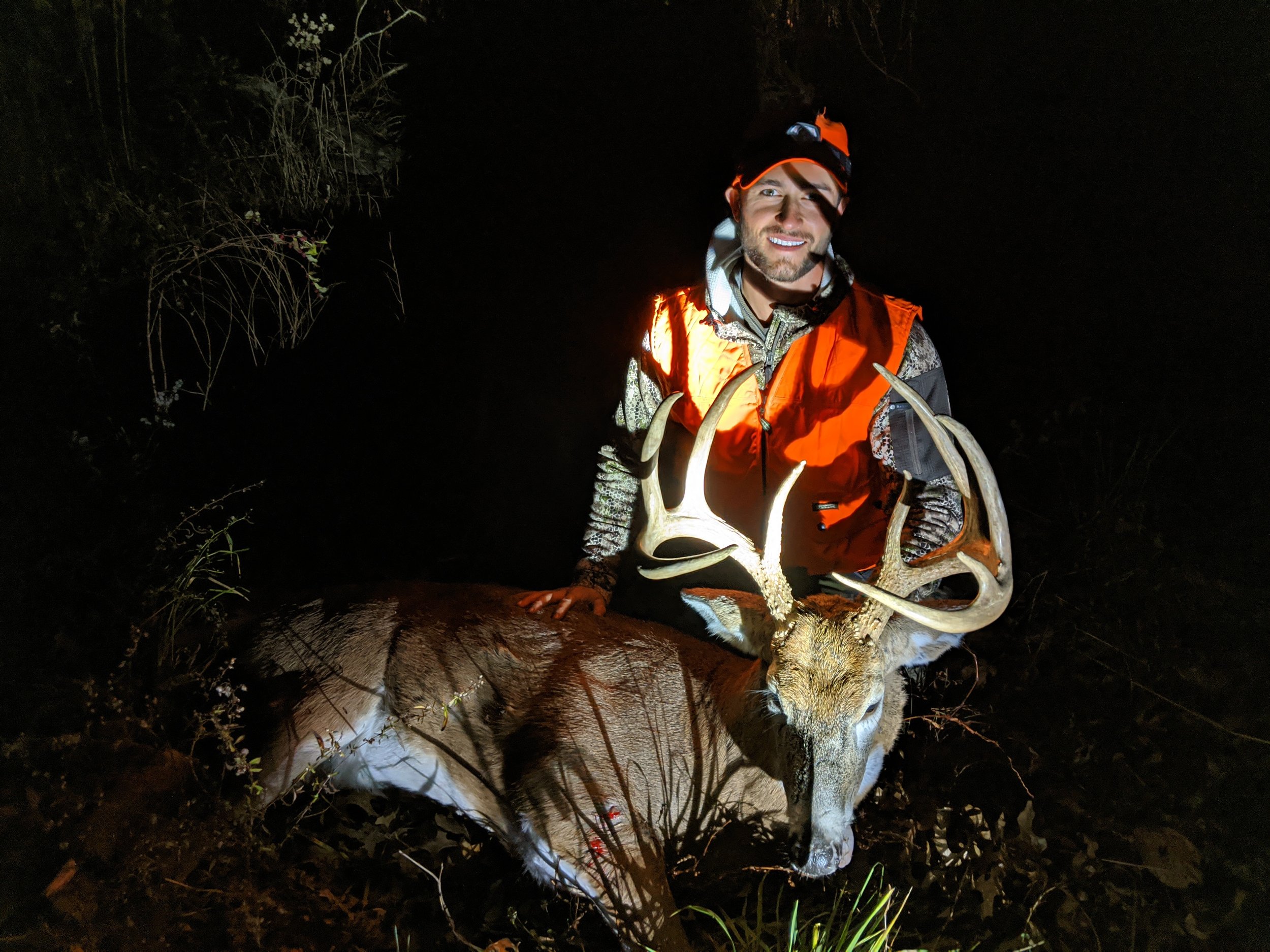 A man wearing a camouflage jacket, orange hunting vest, and orange cap at night, holding a large deer with massive antlers.