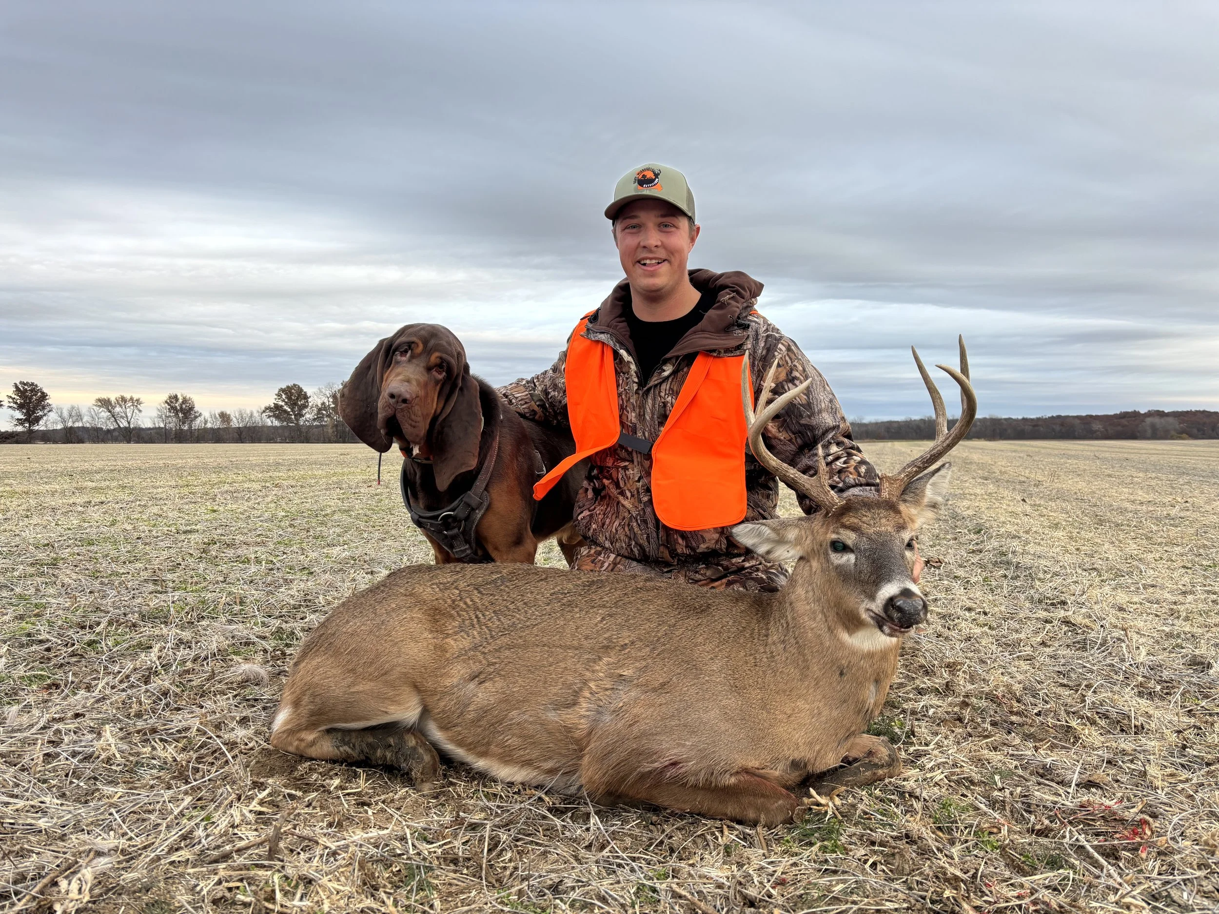 A man kneeling on the ground outdoors wearing camouflage clothing and an orange safety vest, holding a deer and a hunting dog. The deer has antlers and the dog is a brown bloodhound. The background shows a flat field with trees under a cloudy sky.