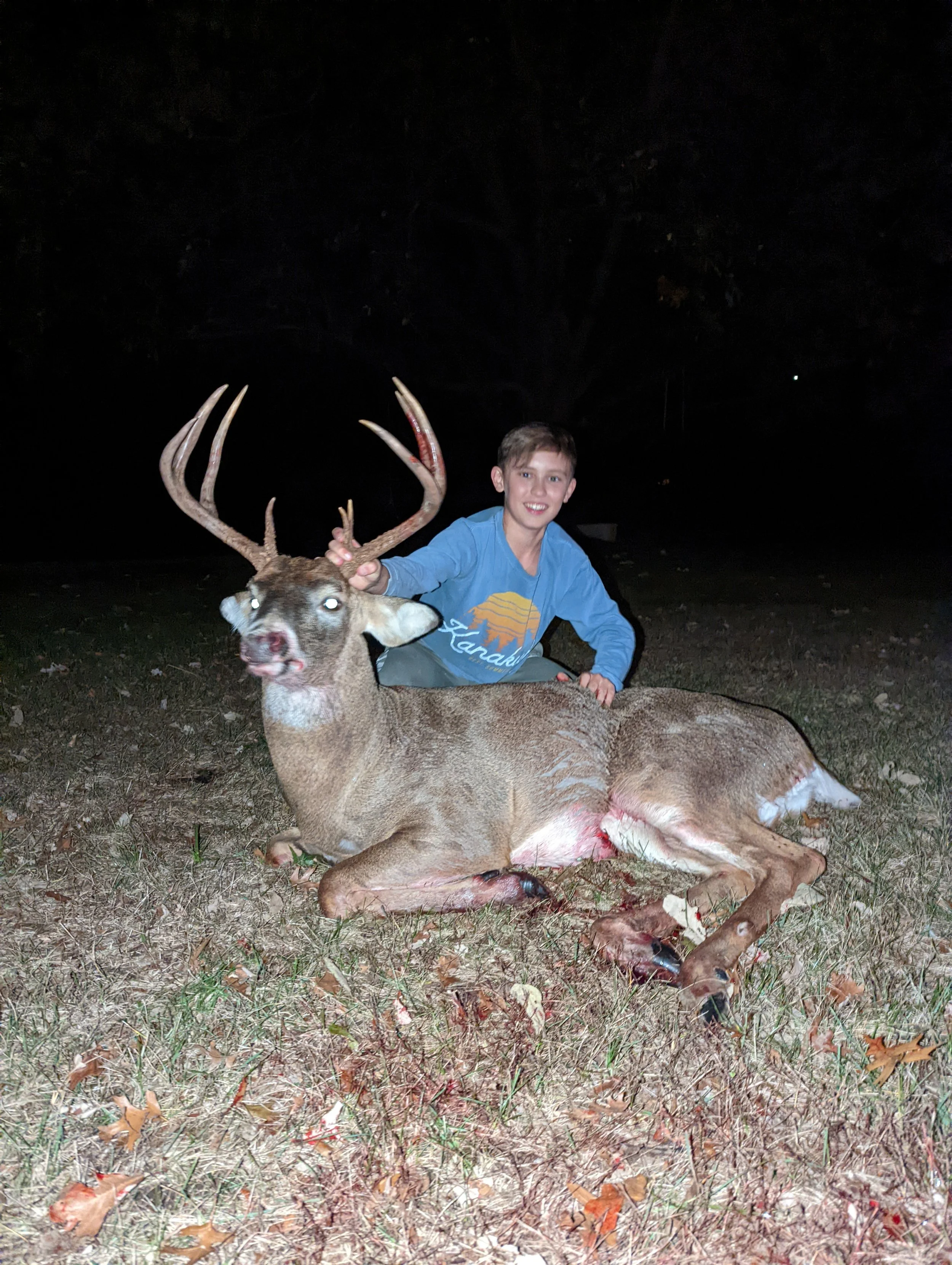A young boy kneeling next to a dead deer with large antlers at night outside, with thatch leaves on the ground and dark trees in the background.