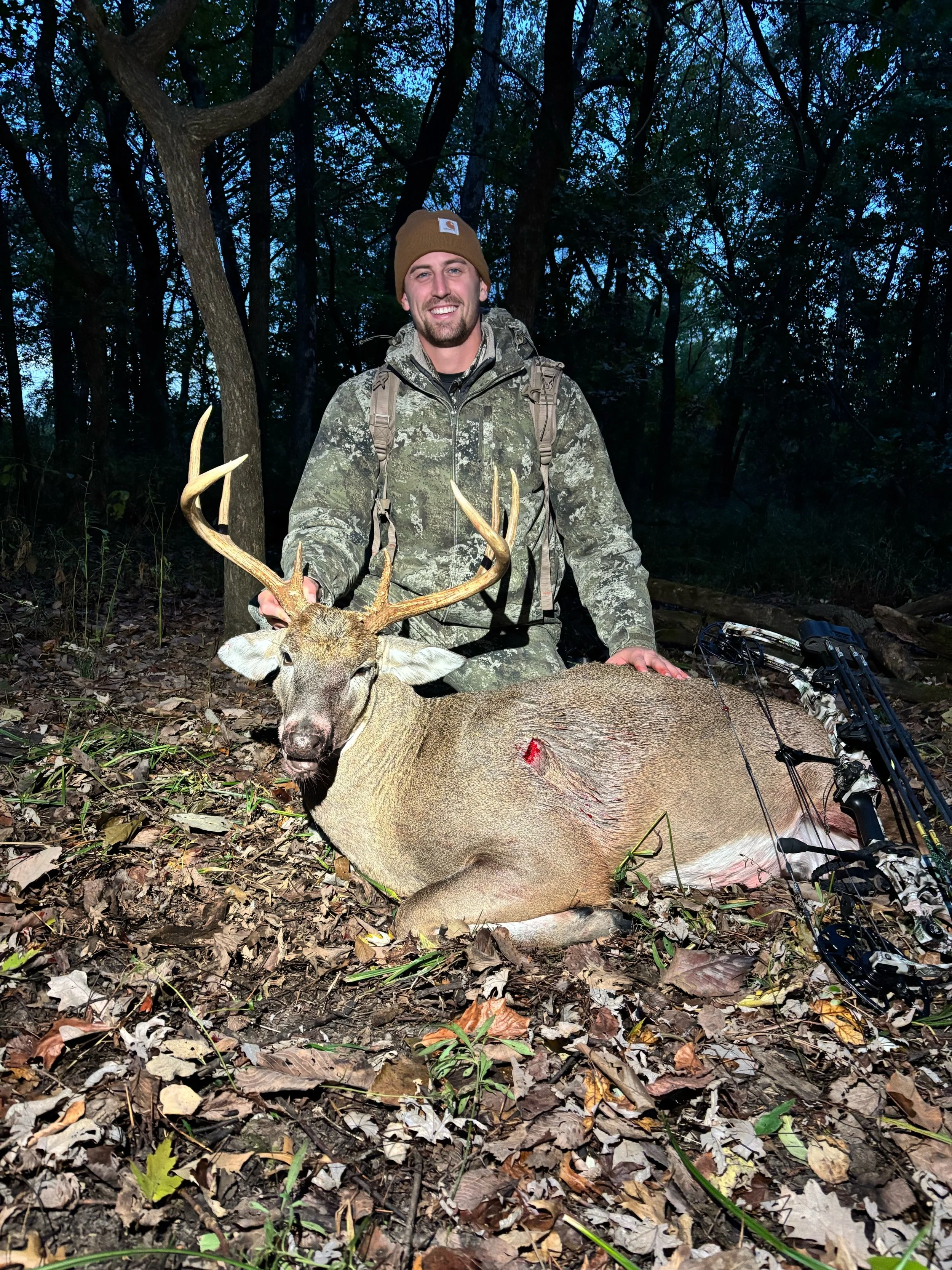 A man in camouflage hunting gear sitting in a forest at dusk, proudly holding a large deer with antlers, a small wound visible on its side.