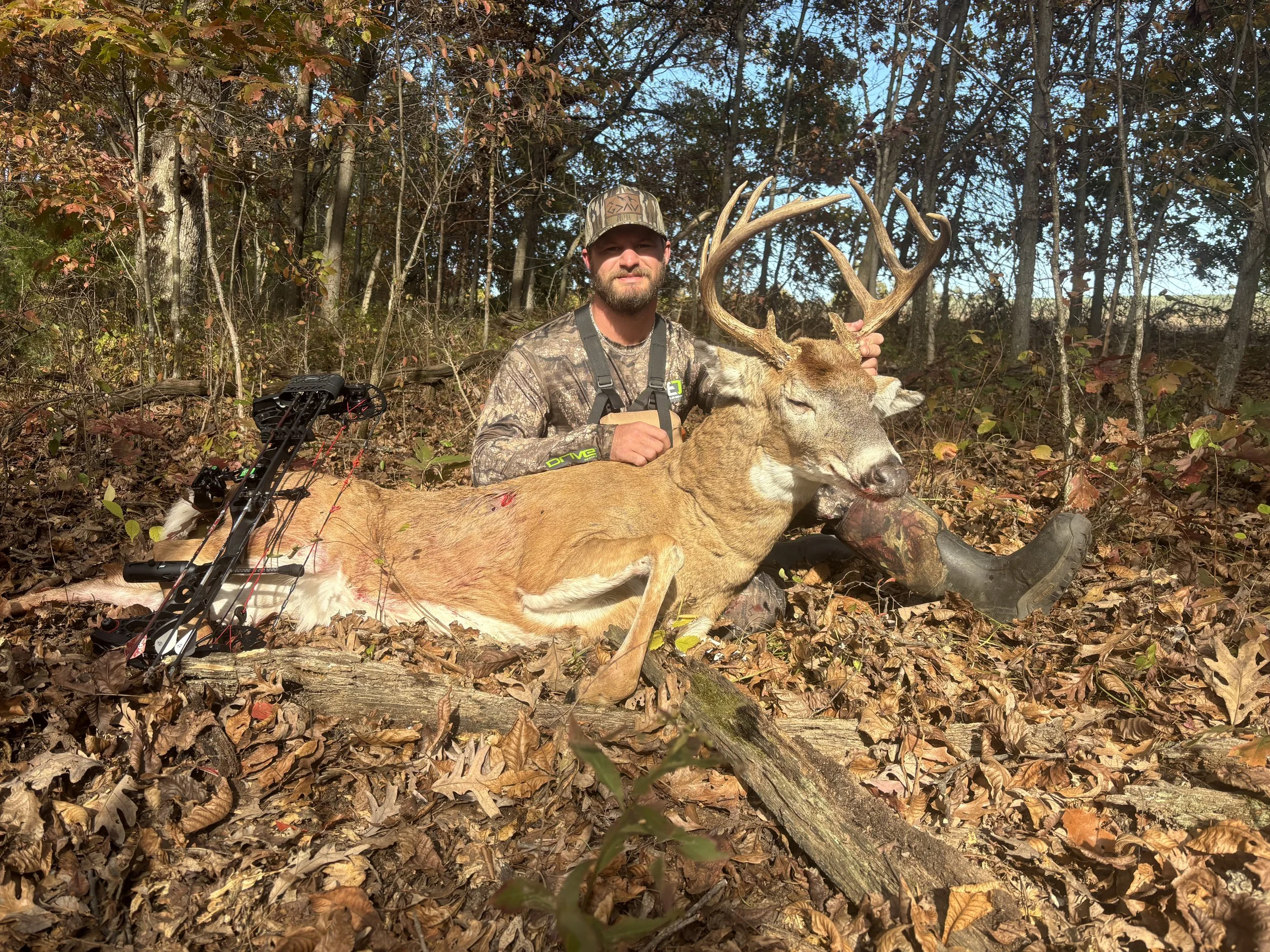A man in camouflage clothing and a cap holding a large deer with antlers. The man is sitting on the ground in a wooded area covered with dried leaves.