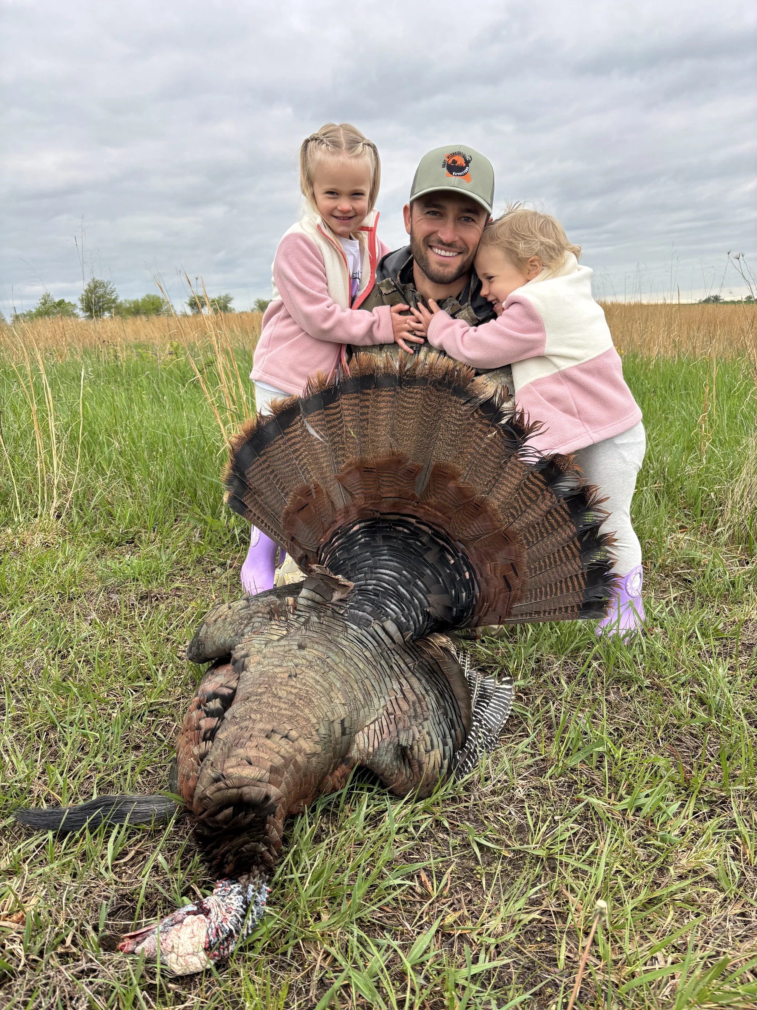 A man and two young girls in a field hold a large turkey with its feathers fanned out. The man is smiling and wearing a cap, while the girls embrace him and smile. The sky is cloudy, and there are tall grass and trees in the background.