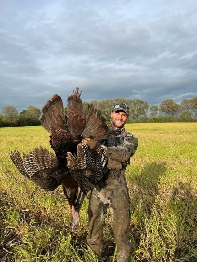 A man dressed in camouflage clothing standing in a grassy field, holding a large turkey with both hands after a successful hunt, with trees and a cloudy sky in the background.