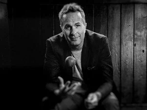 Black and white photo of a man sitting indoors, wearing a suit jacket, casually tossing a ball. Wooden wall background, low lighting.