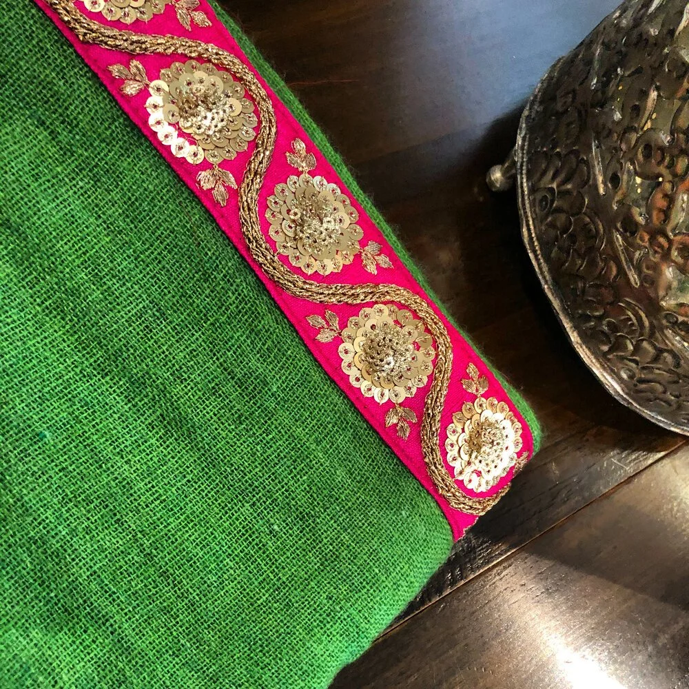 Close-up of a green fabric with a pink and gold embroidered border. Part of a metallic decorative object is visible on the right.