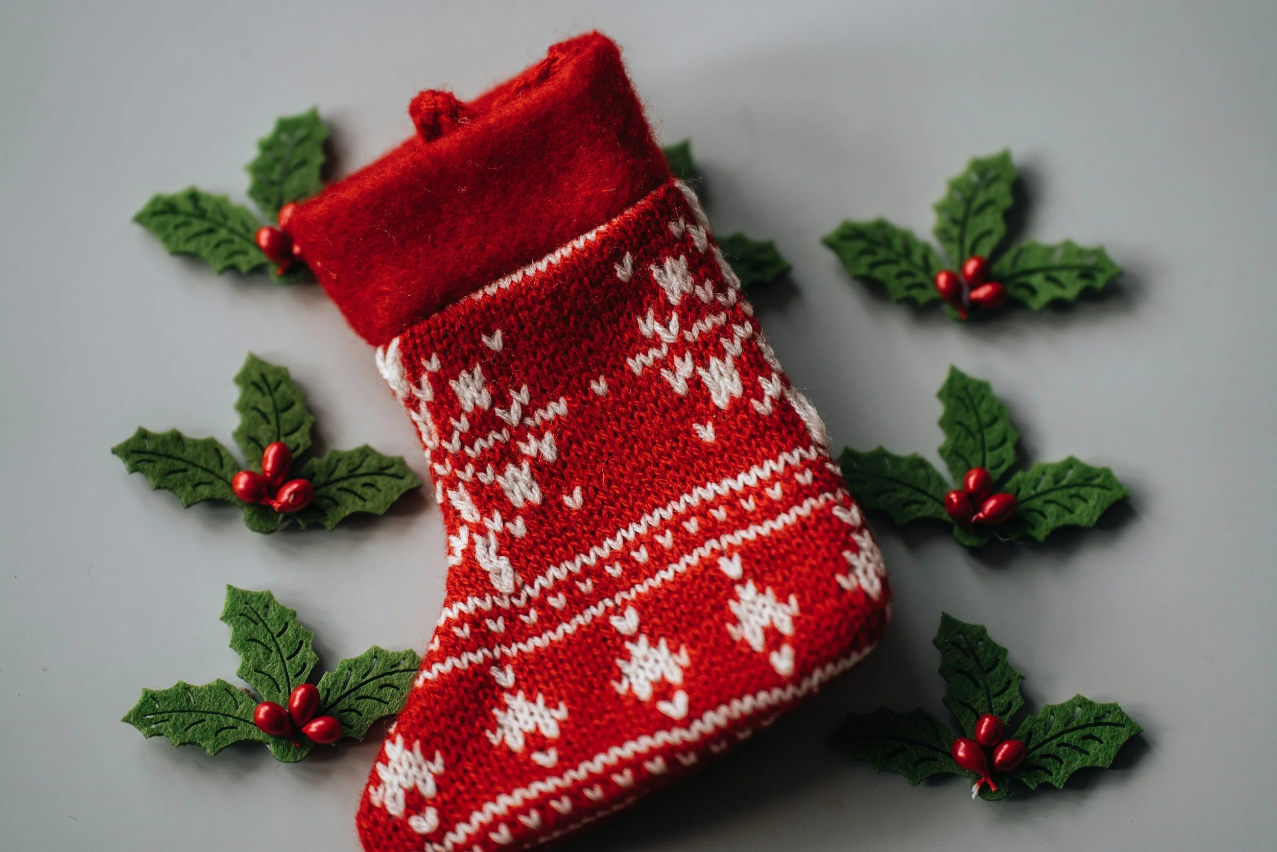 A red Christmas stocking with white snowflake and heart patterns surrounded by holly leaves with red berries on a light background.