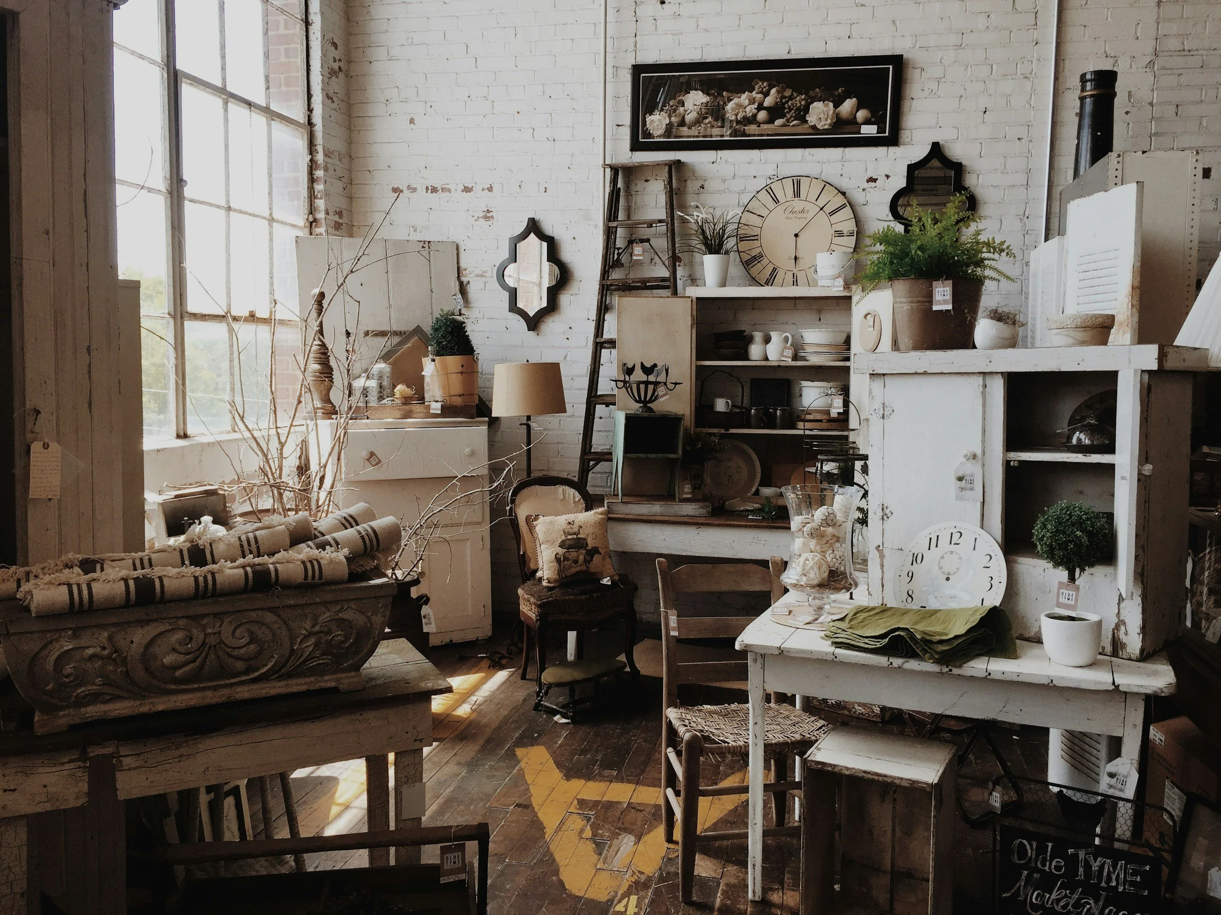 Interior of a vintage-style store with white brick walls, L-shaped shelving, a clock, framed pictures, plants, and furniture, including a chandelier, chairs, and a table with greenery and decorative items.