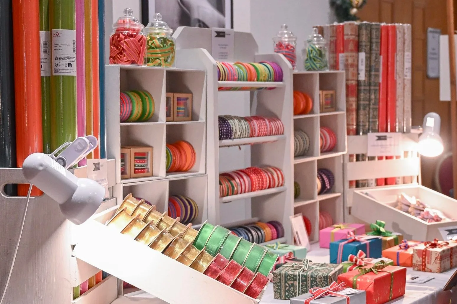 A woman shopping at a holiday gift booth with wrapped presents and colorful ribbon rolls, Christmas decorations, and boxes for sale, while looking at her phone.