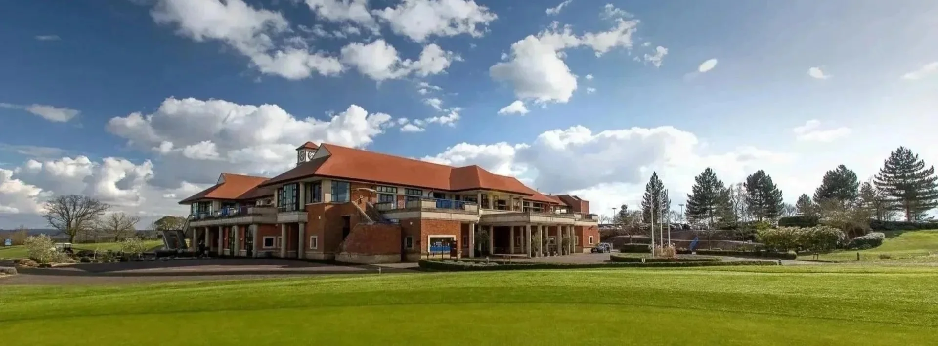 Large building with a red-tiled roof, surrounded by a grassy area and trees, under a partly cloudy sky.