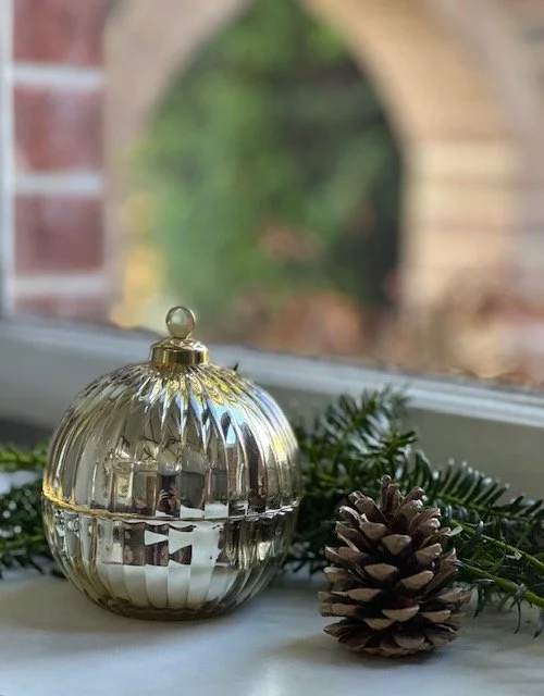 A decorative glass ornament with gold accents, a pinecone, and pine branches on a windowsill with a blurry outdoor view.