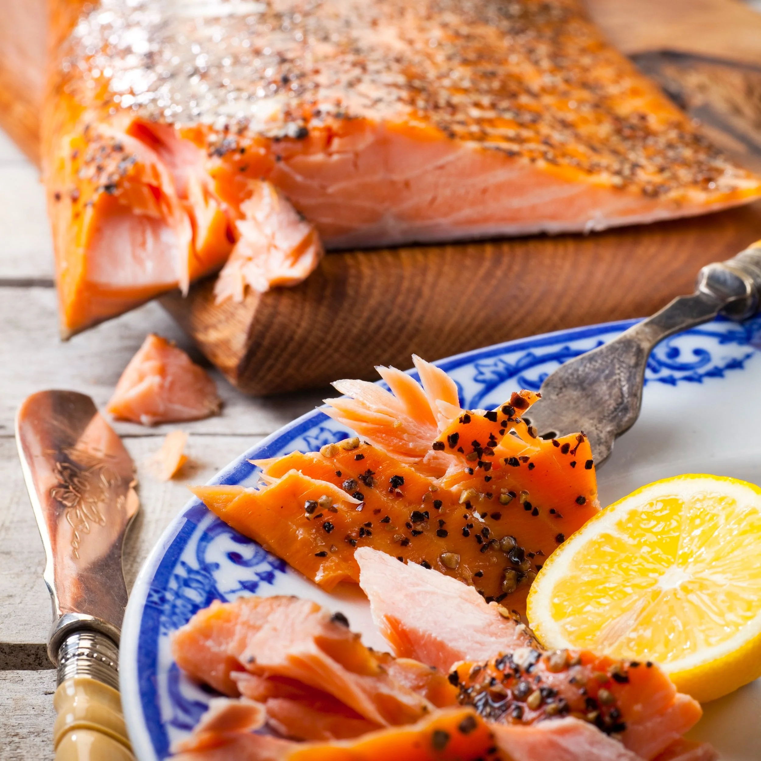 Close-up of sliced smoked salmon with black pepper and lemon slices on a blue and white plate, with a large fillet of smoked salmon in the background.