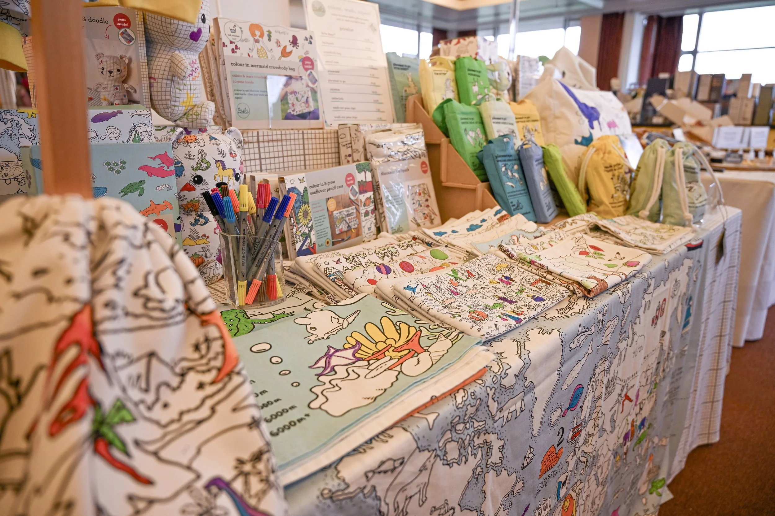 A display table with coloring books, crossbody bags, and art supplies at an indoor craft fair.