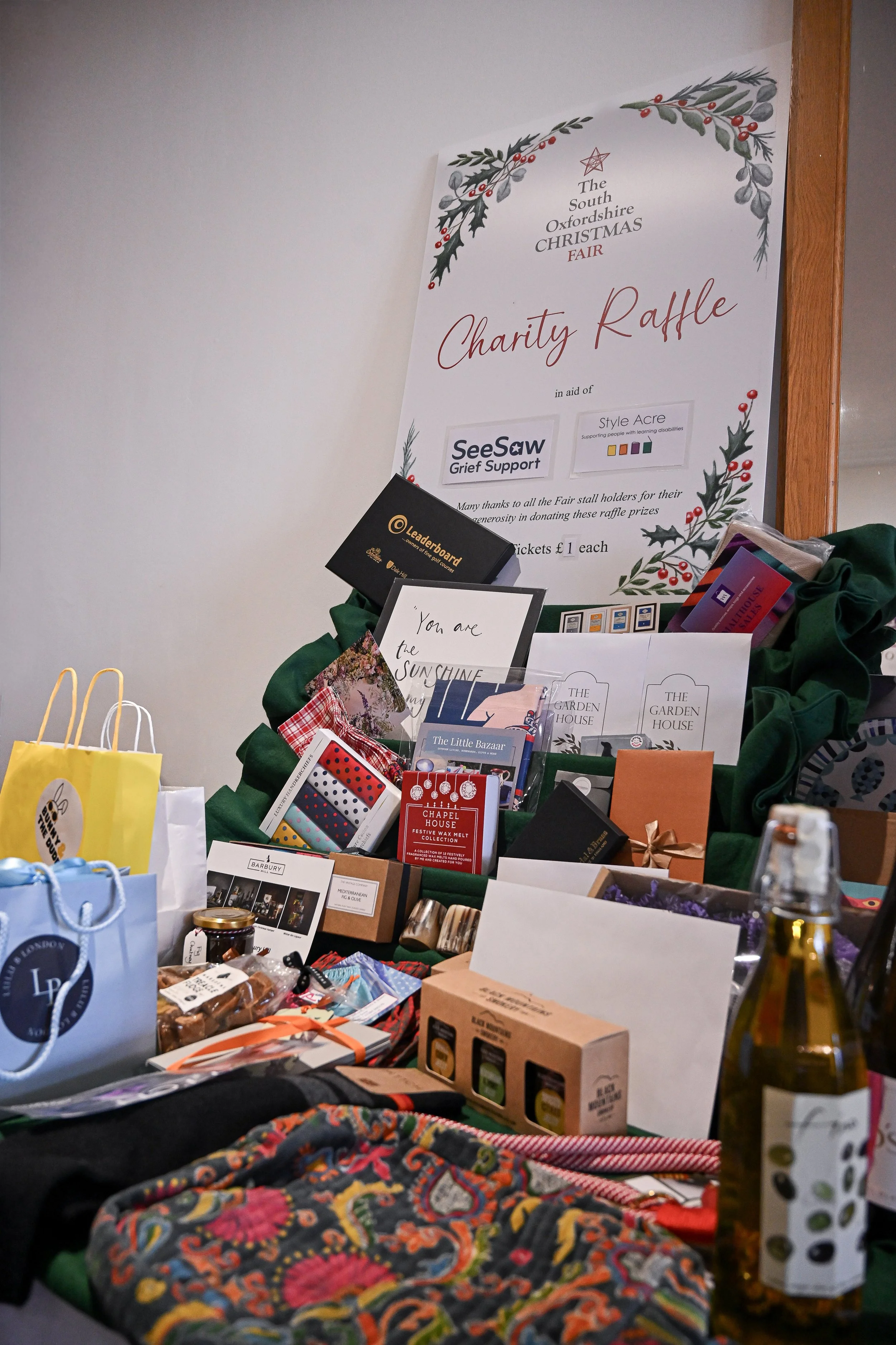 A charity raffle display table at a Christmas fair with various wrapped gifts, items, and promotional materials, set against a white wall with a large poster for the event.