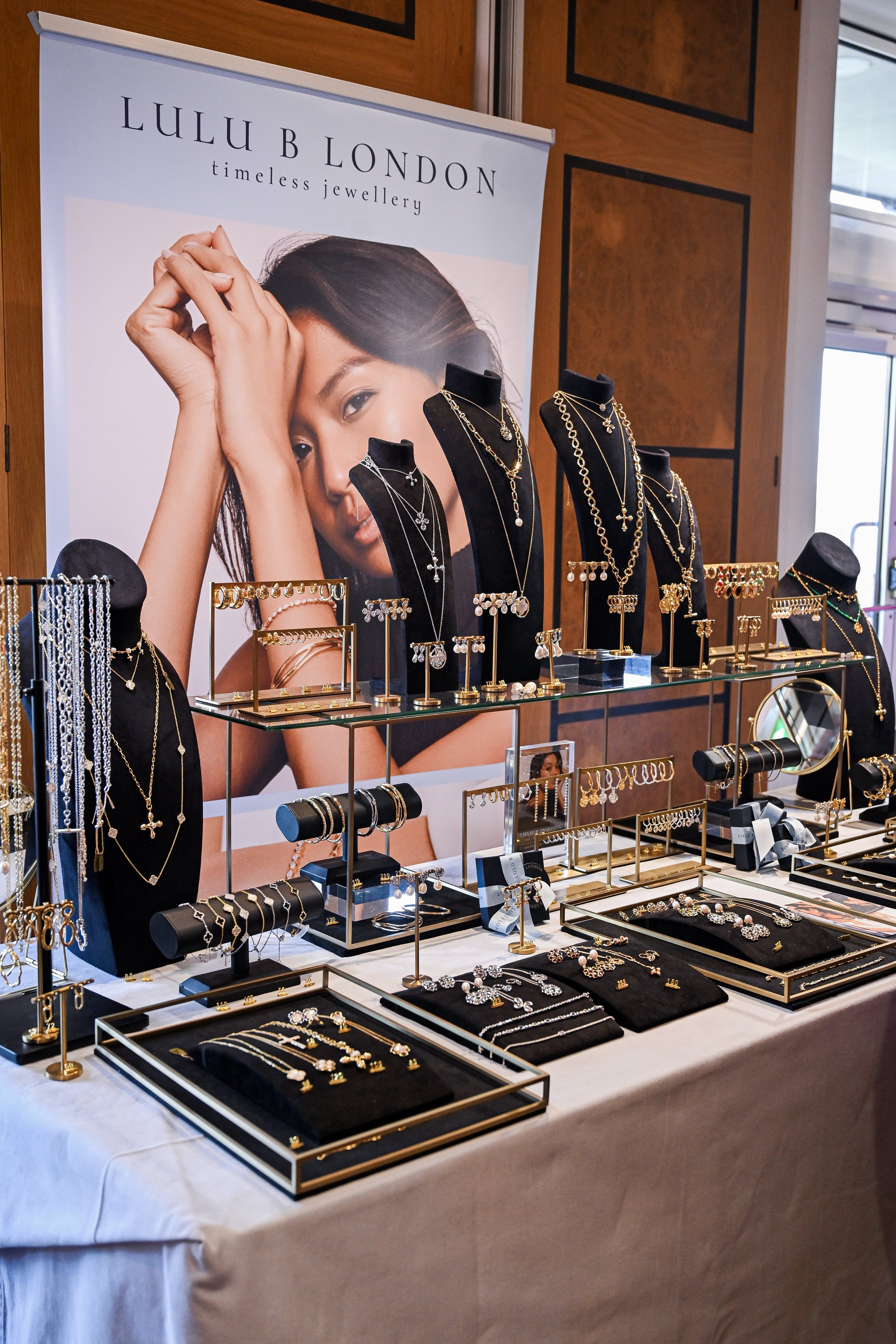Jewelry display at a Lulu B London booth, showcasing necklaces, earrings, bracelets, and rings with a large poster of a woman modeling jewelry in the background.