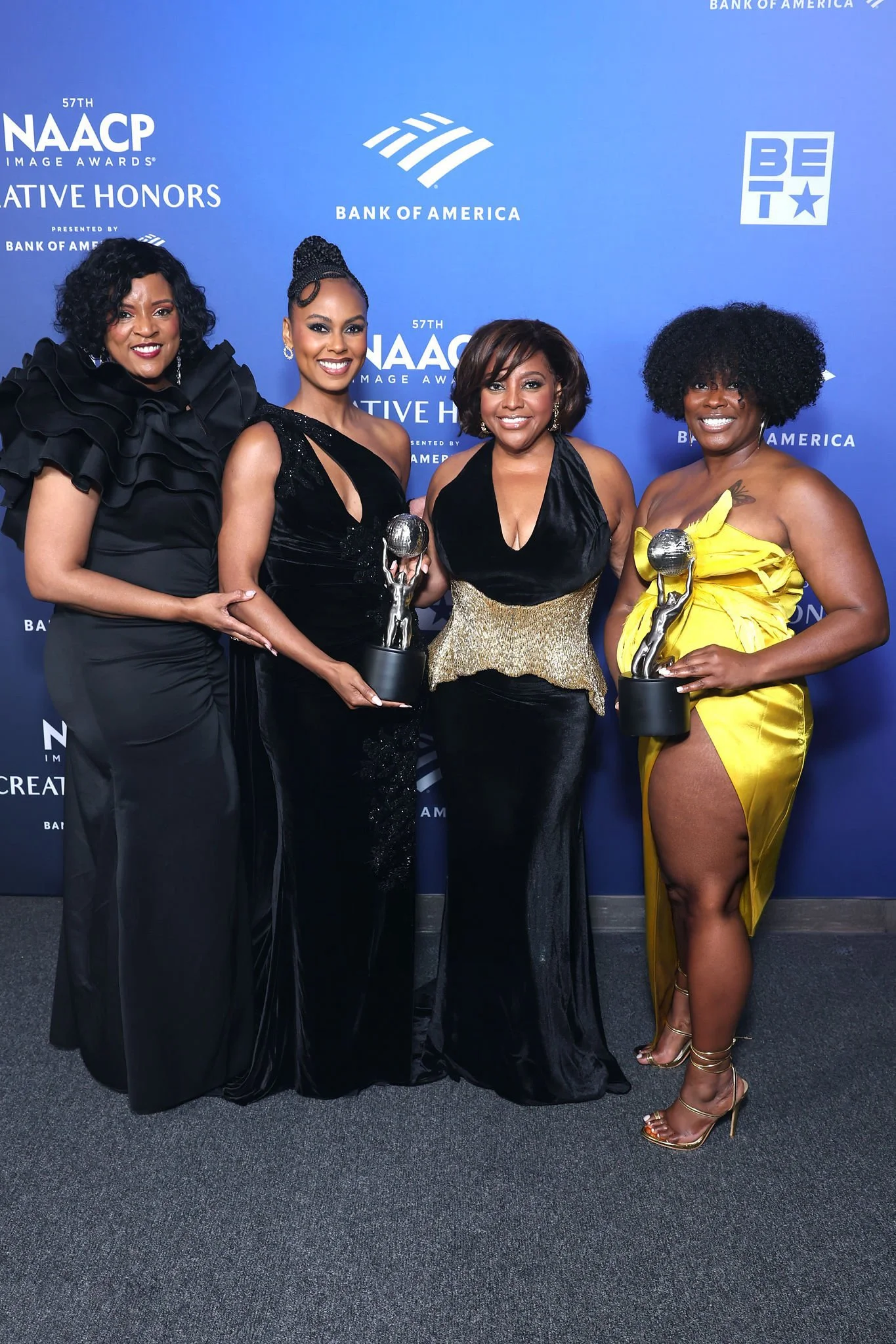 Ashley Versher in a Tess Mann Atelier gown. Four women in elegant dresses standing together at the NAACP Image Awards, holding trophies, against a blue backdrop featuring the NAACP logo and sponsors.