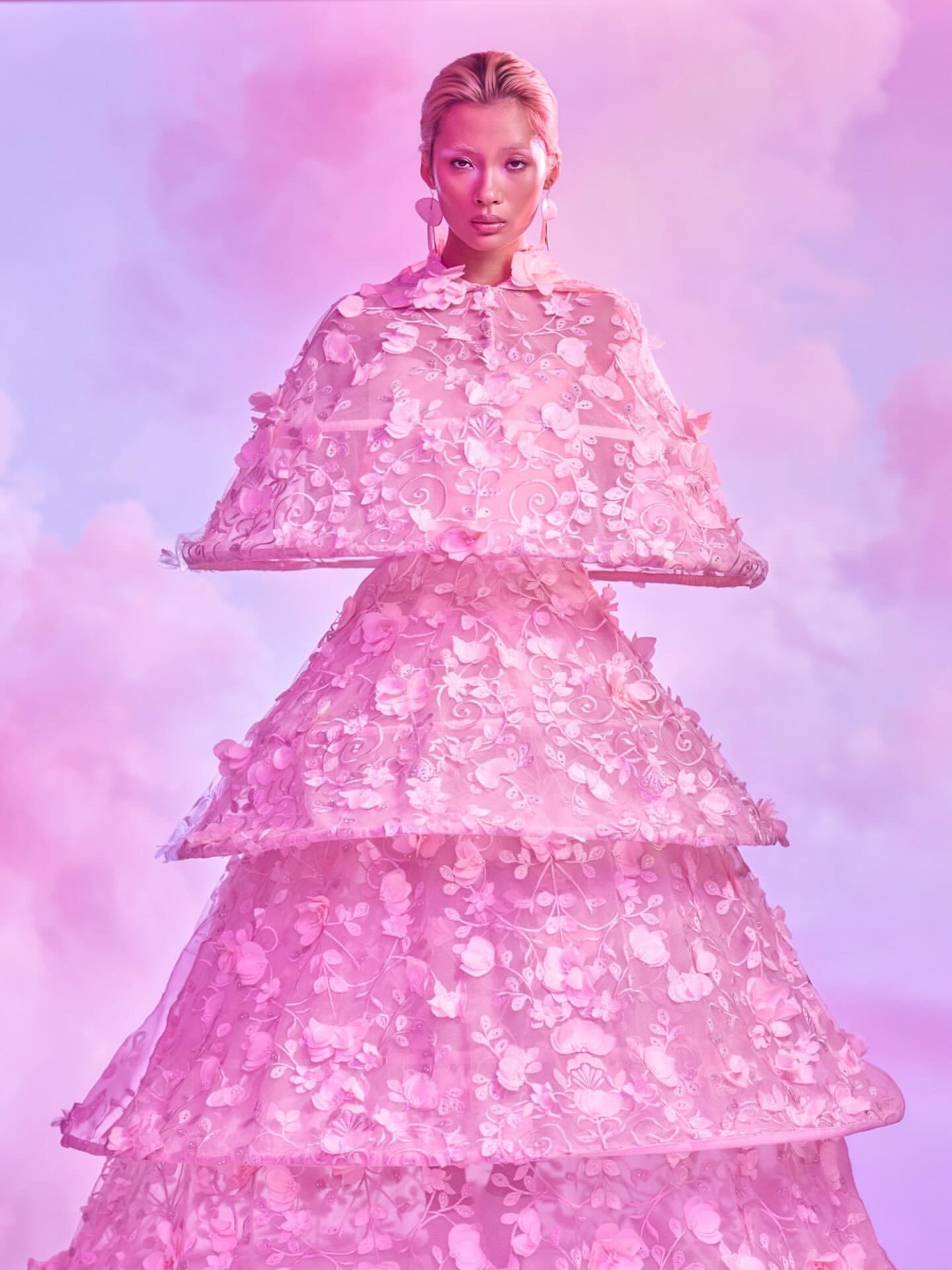 Woman in a pink, floral-tiered dress with a matching wide-brimmed hat against a pastel pink and purple cloudy background. Tess Mann Atelier by Lindsay Adler for Canon