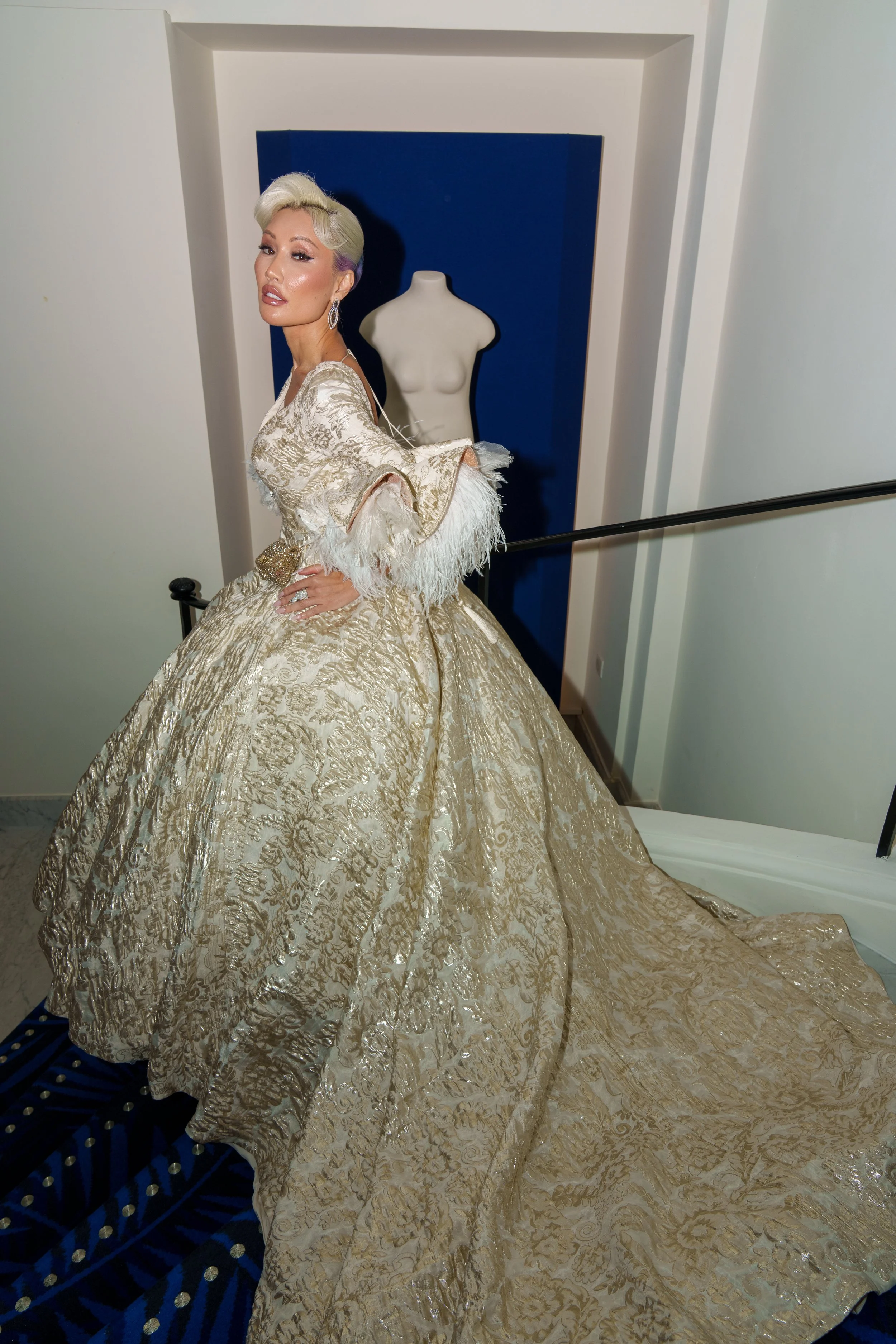 Aliia Roza in an ornate gold and cream Tess Mann Atelier gown with feathered sleeves, standing on a staircase in front of a blue background with a mannequin torso in the background at Cannes Film Festival.