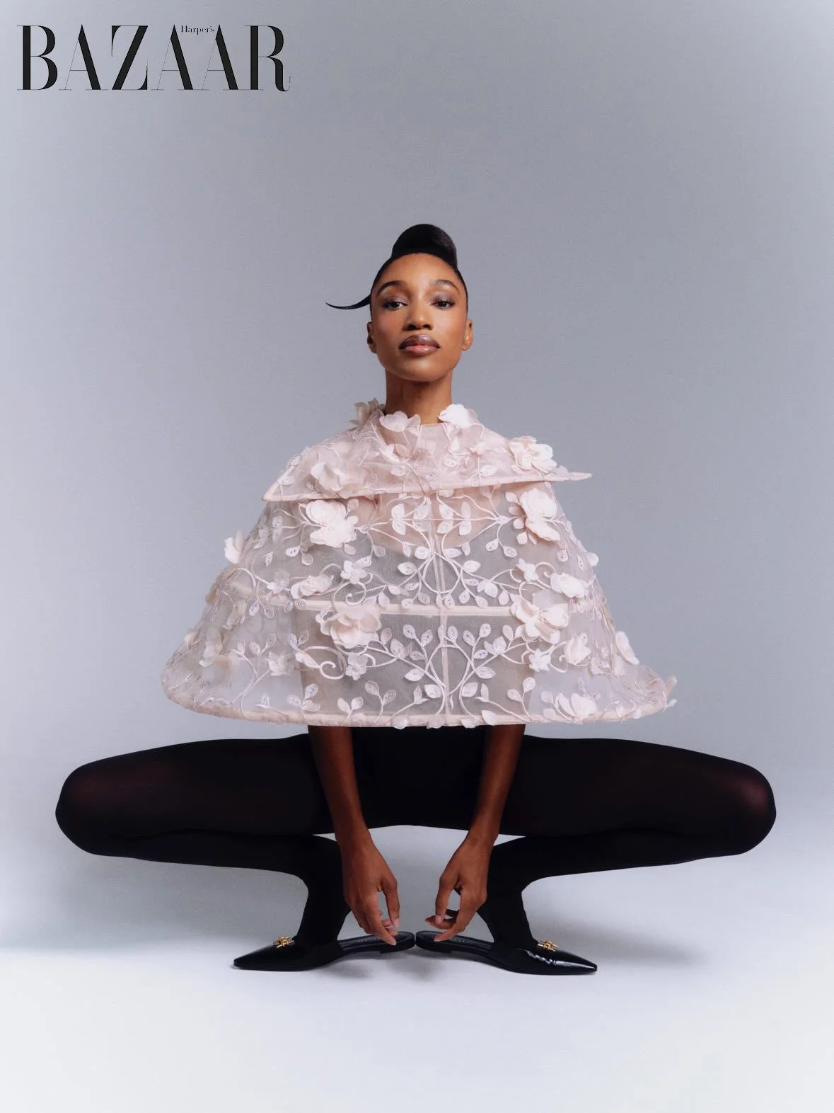 A fashion magazine cover featuring a woman in an avant-garde, floral lace cape, black tights, and black pointed shoes with gold embellishments, posed in a squatting position against a plain gray background, with the 'Harper's Bazaar' logo at the top.